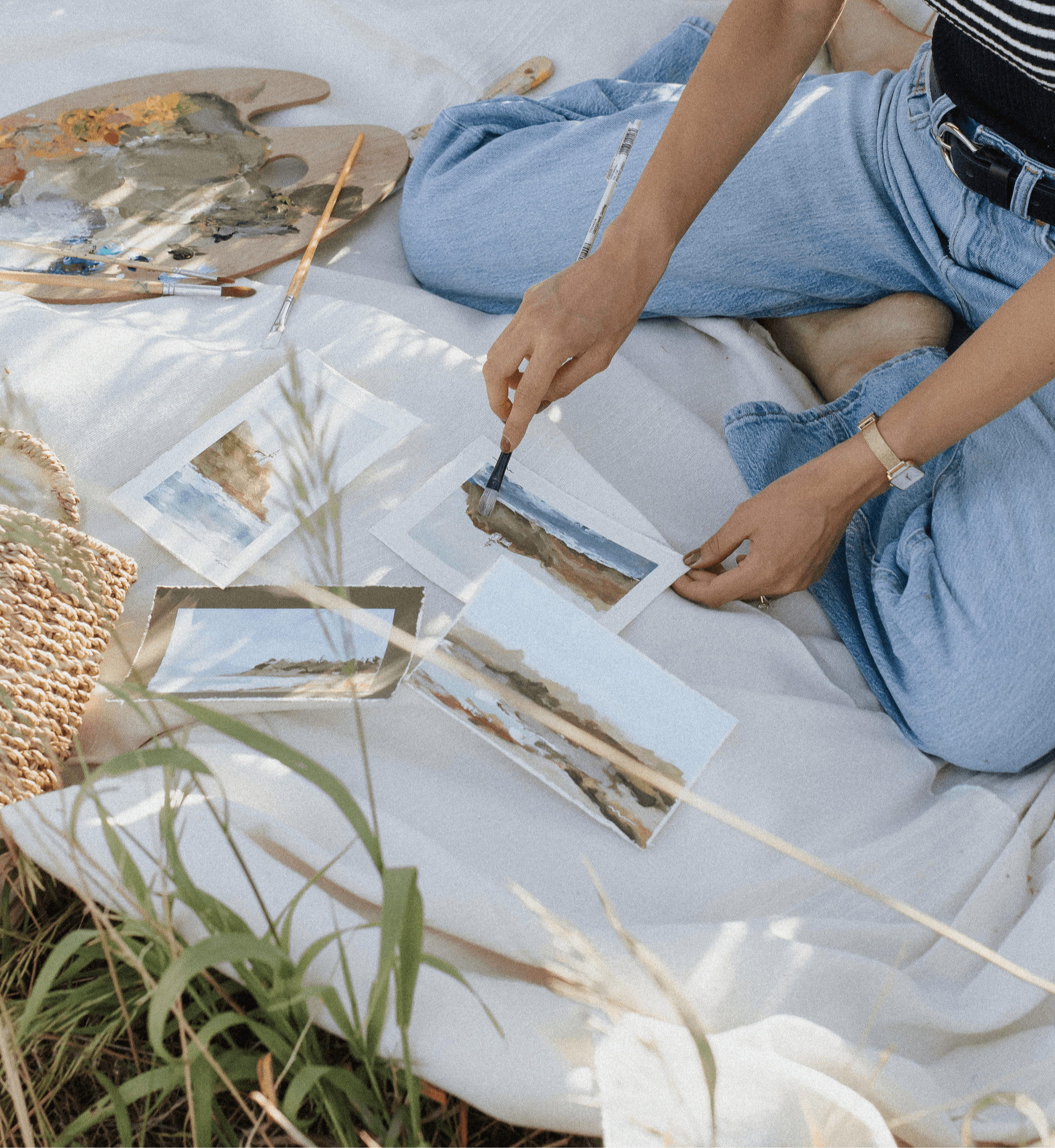 Person sitting on a white cloth, arranging photographs of landscapes, with painting supplies nearby, outdoors. Word count: 20.
