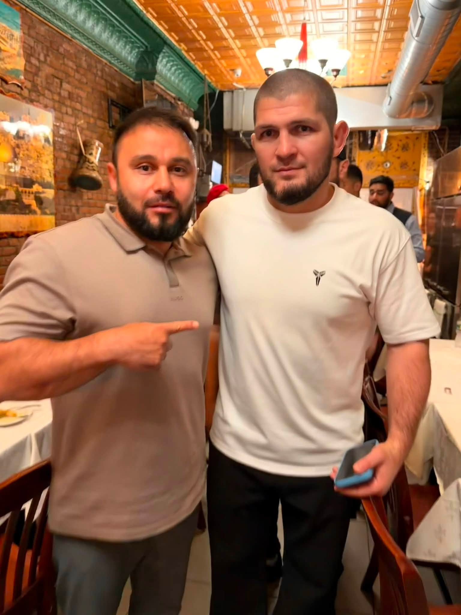 Two men standing together in a restaurant, one pointing at the other, with warm lighting and brick walls in the background.