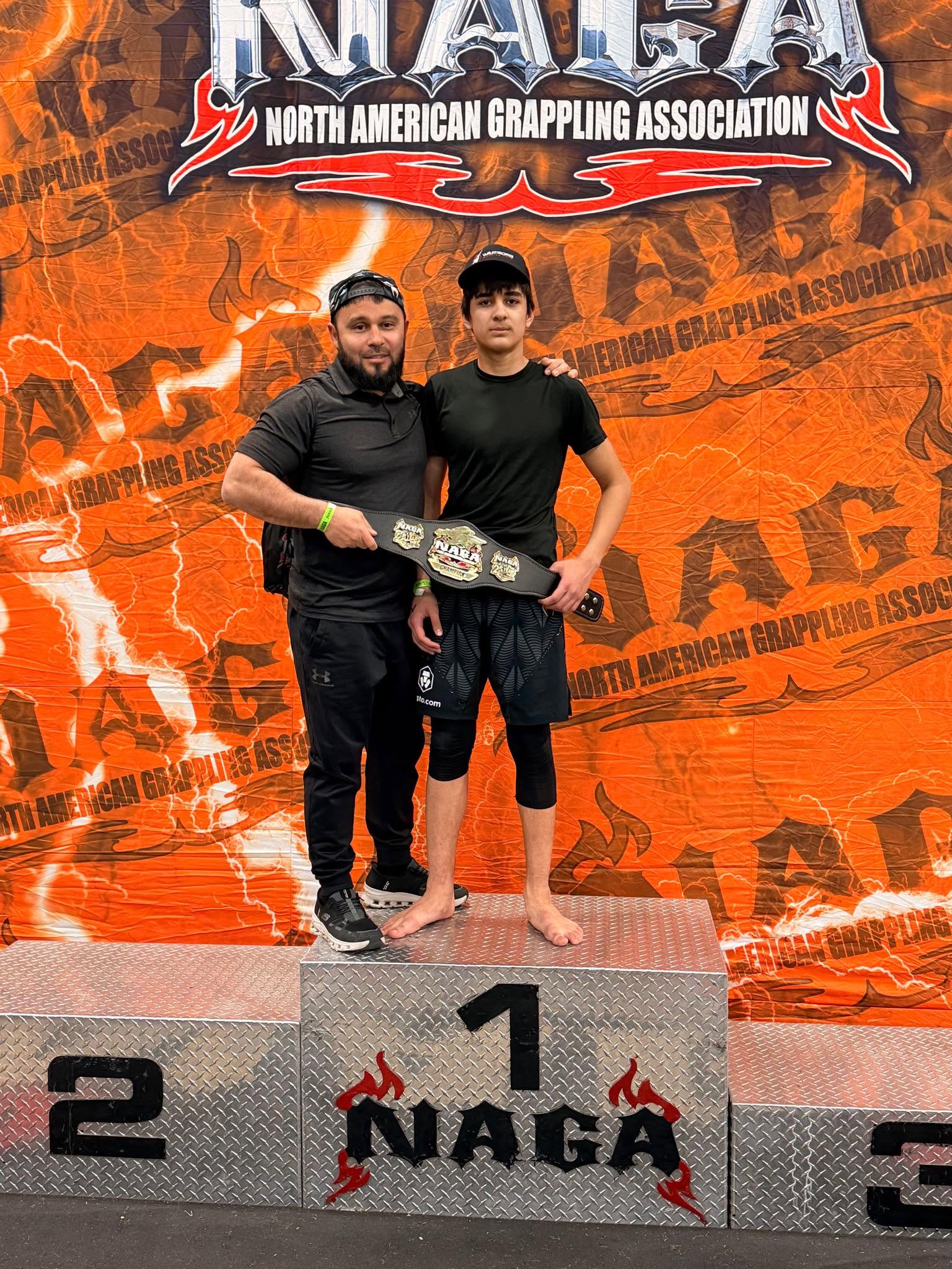 Two men stand on a winners' podium, one holding a championship belt, with an orange backdrop and NAGA logo.