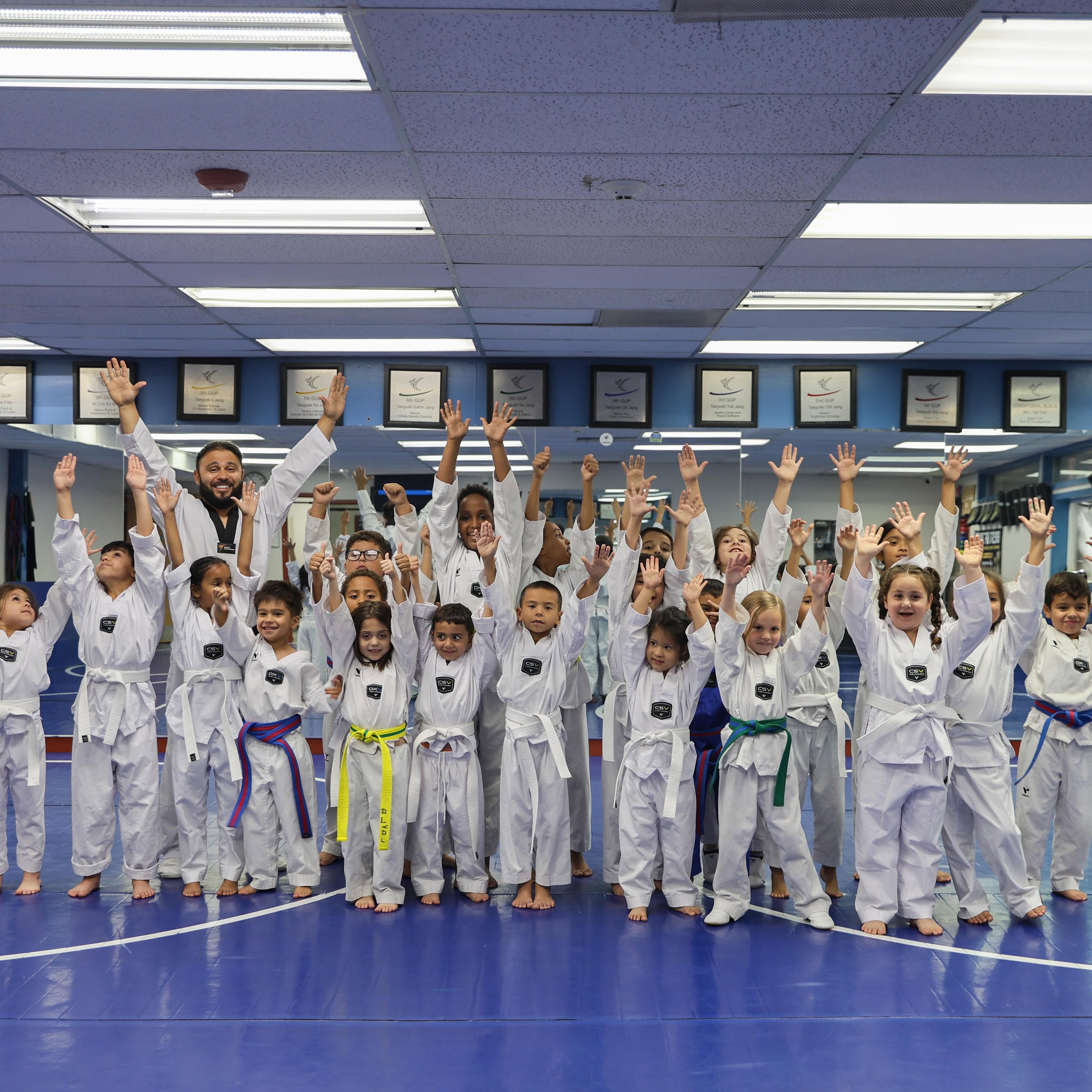 Group of children and instructor in martial arts uniforms raising hands in a dojo, blue floor, ceiling lights, and monitors in background.
