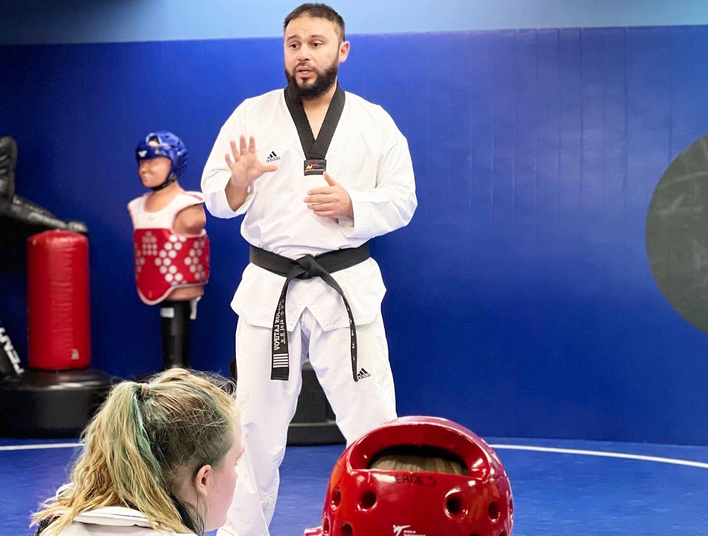 Martial arts instructor in a white gi with a black belt speaking to students in a training room.