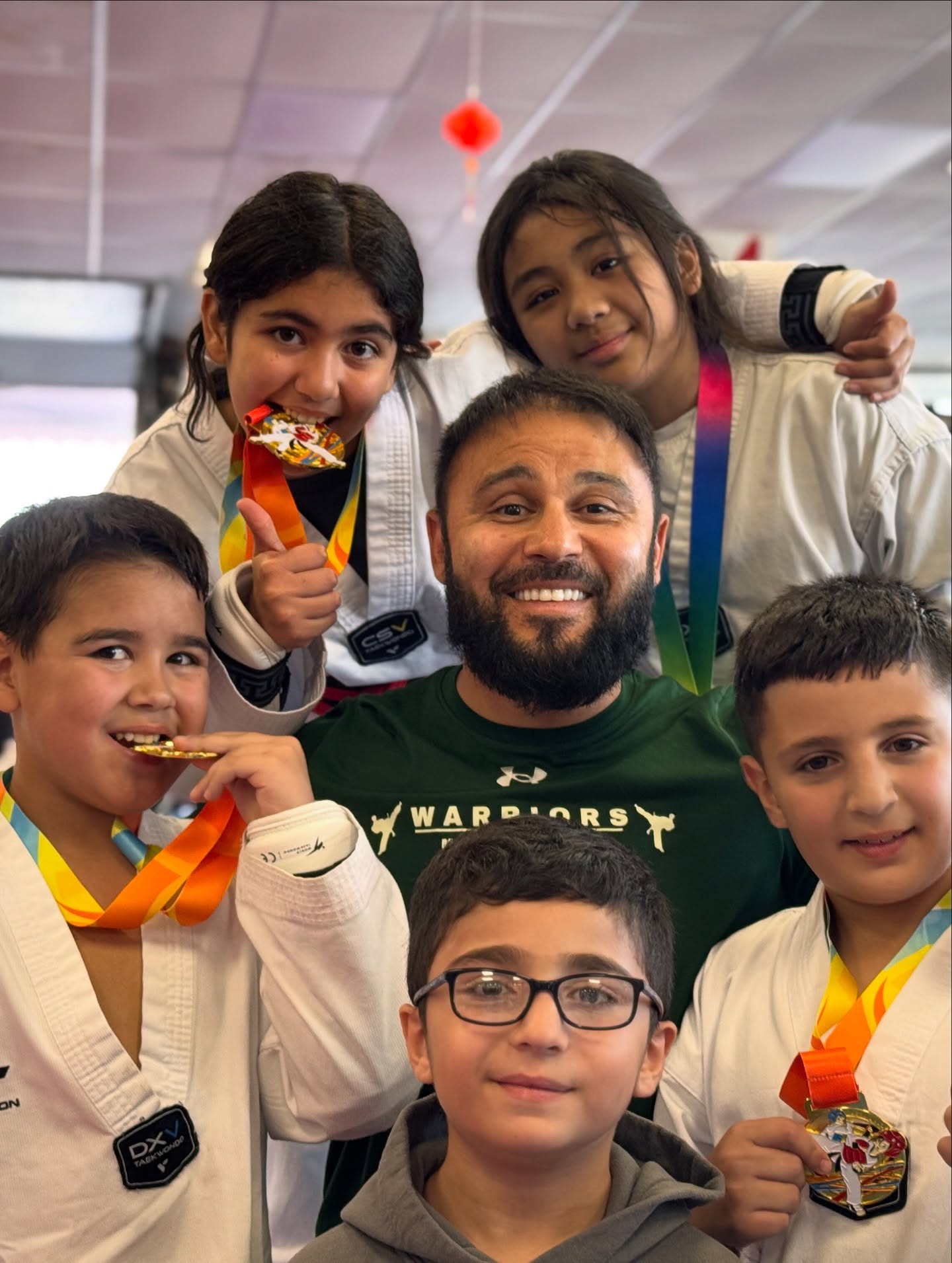 Group of children and a man smiling, wearing martial arts uniforms and medals, indoors with a ceiling and lights visible.