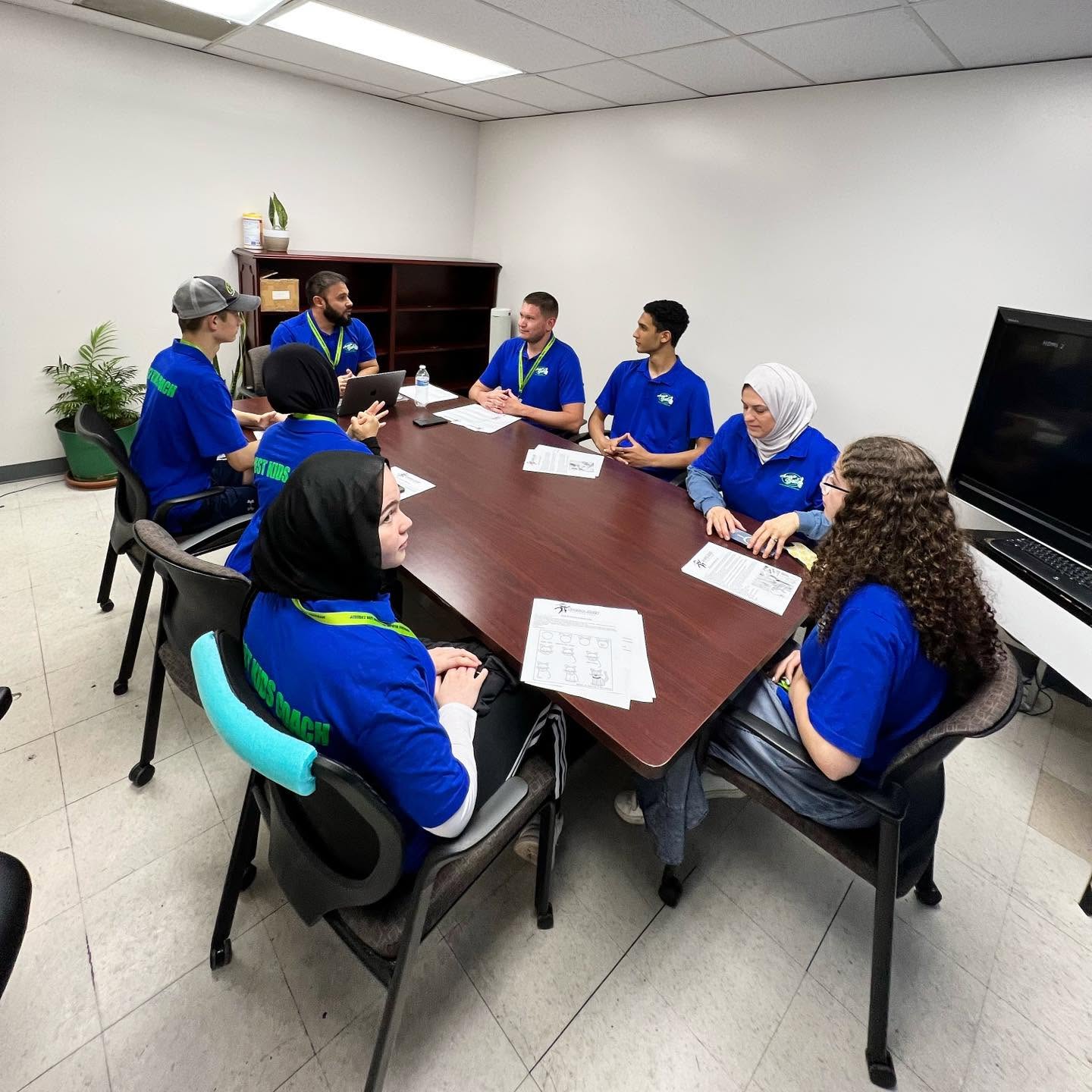 Group of eight people in blue uniforms sitting around a conference table in a room with white walls and a large TV screen.