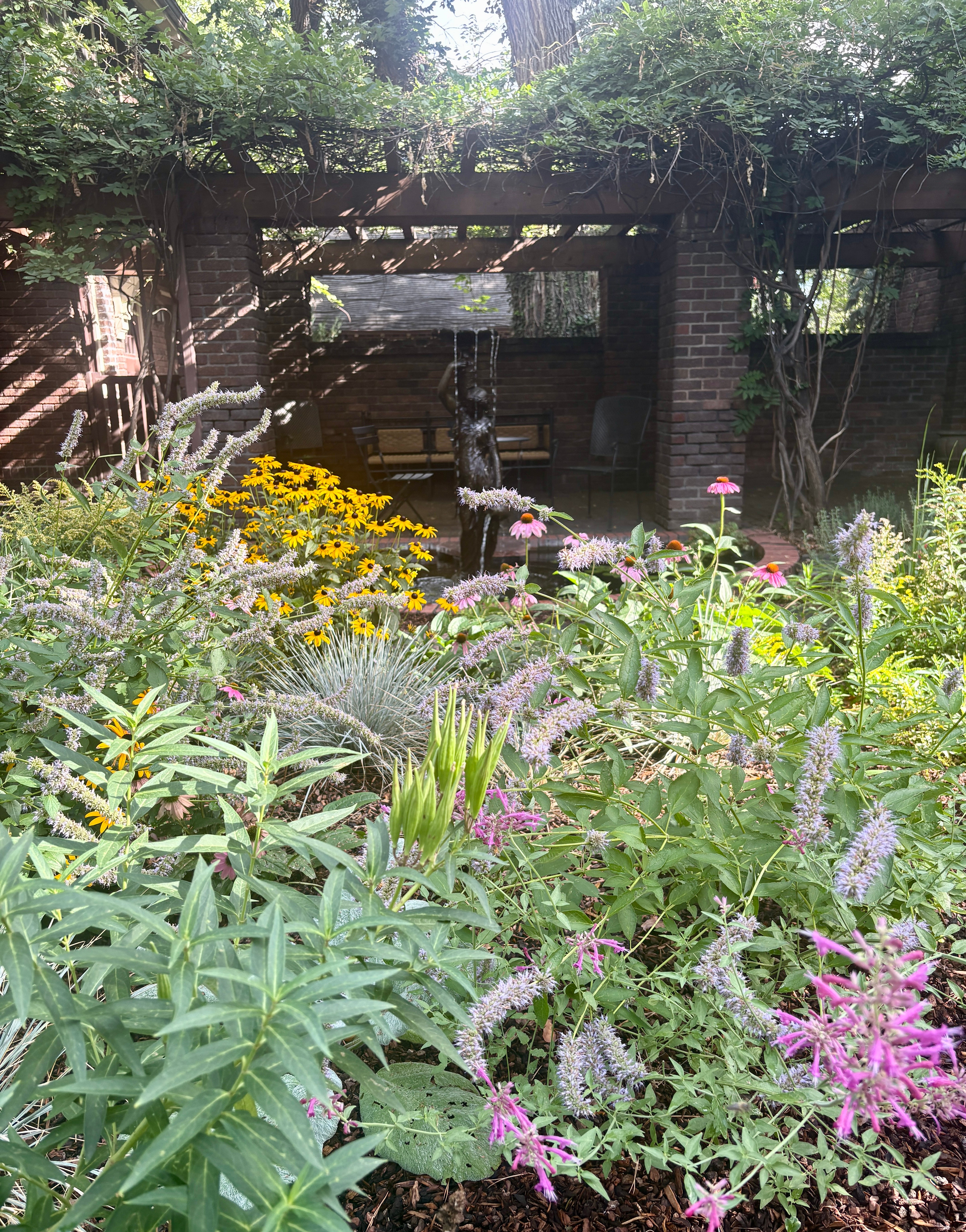 Lush garden with colorful flowers and greenery in front of a brick structure with stairs and a wooden overhang.