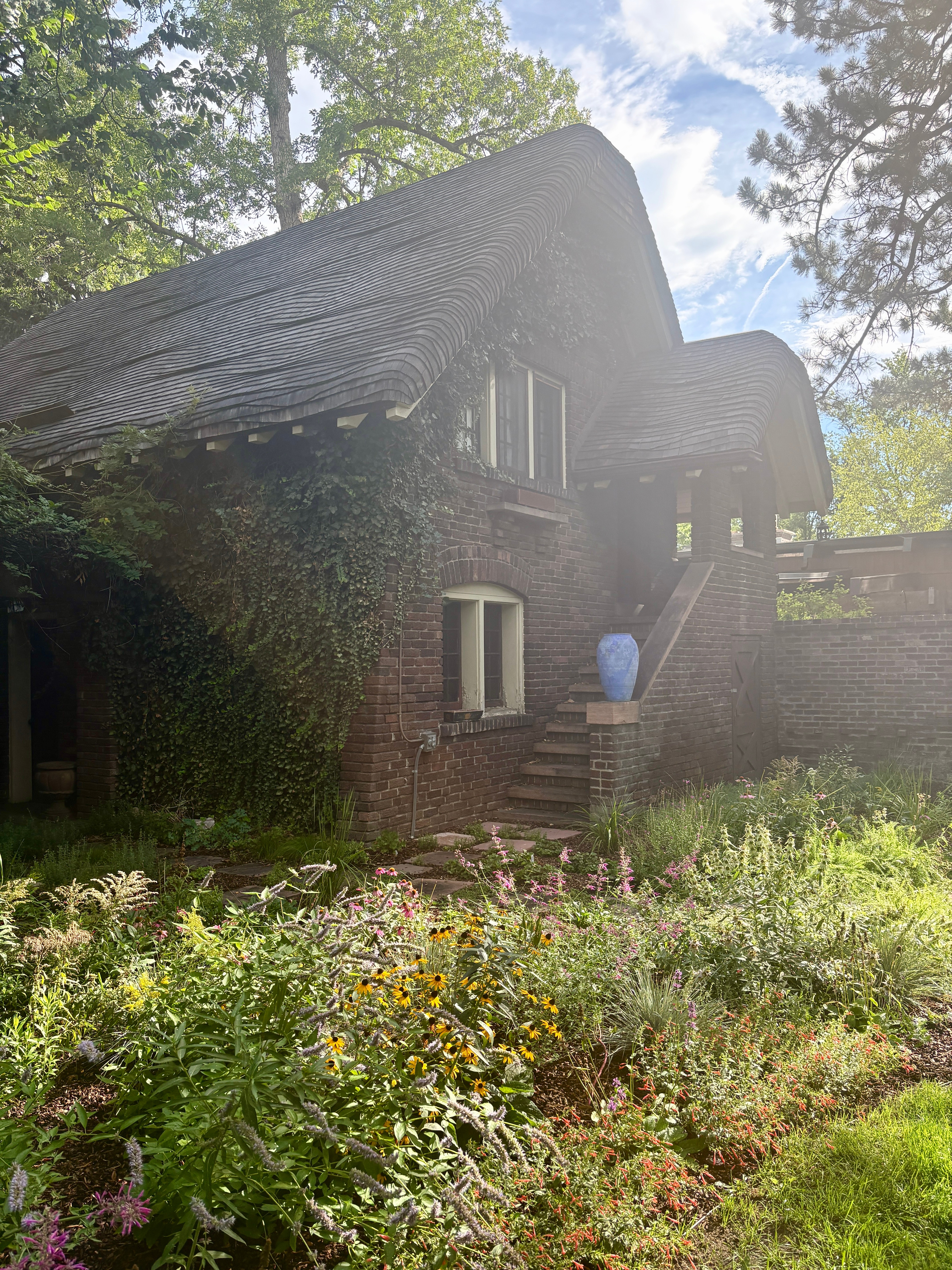 House with a thatched roof, brick walls, surrounded by greenery and flowers, with stairs leading to the entrance.