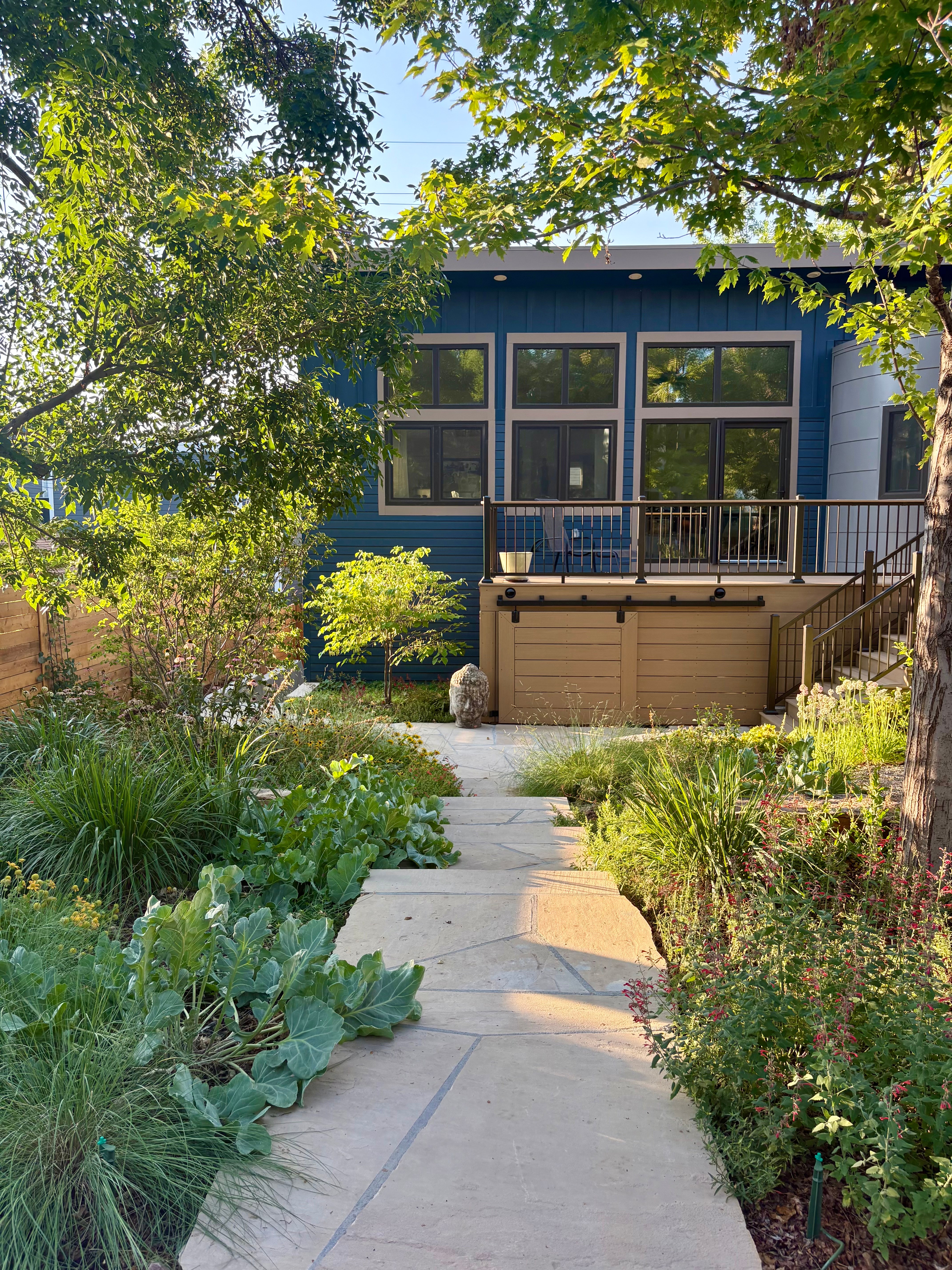 Pathway leading to a house with large windows, surrounded by greenery and trees, in sunlight.