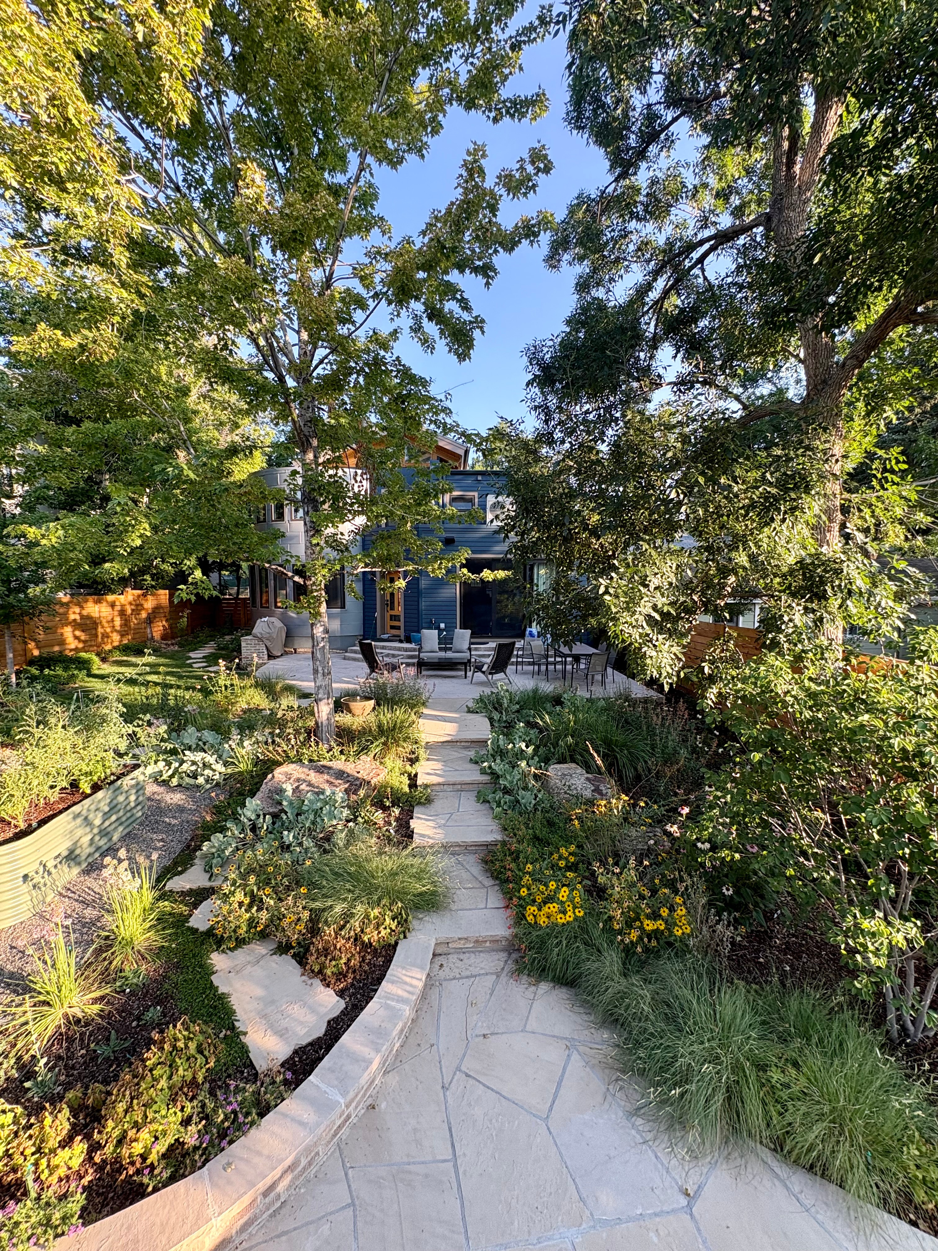 Pathway through a lush garden with trees, plants, and flowers leading to a house with outdoor seating area, sunny sky.