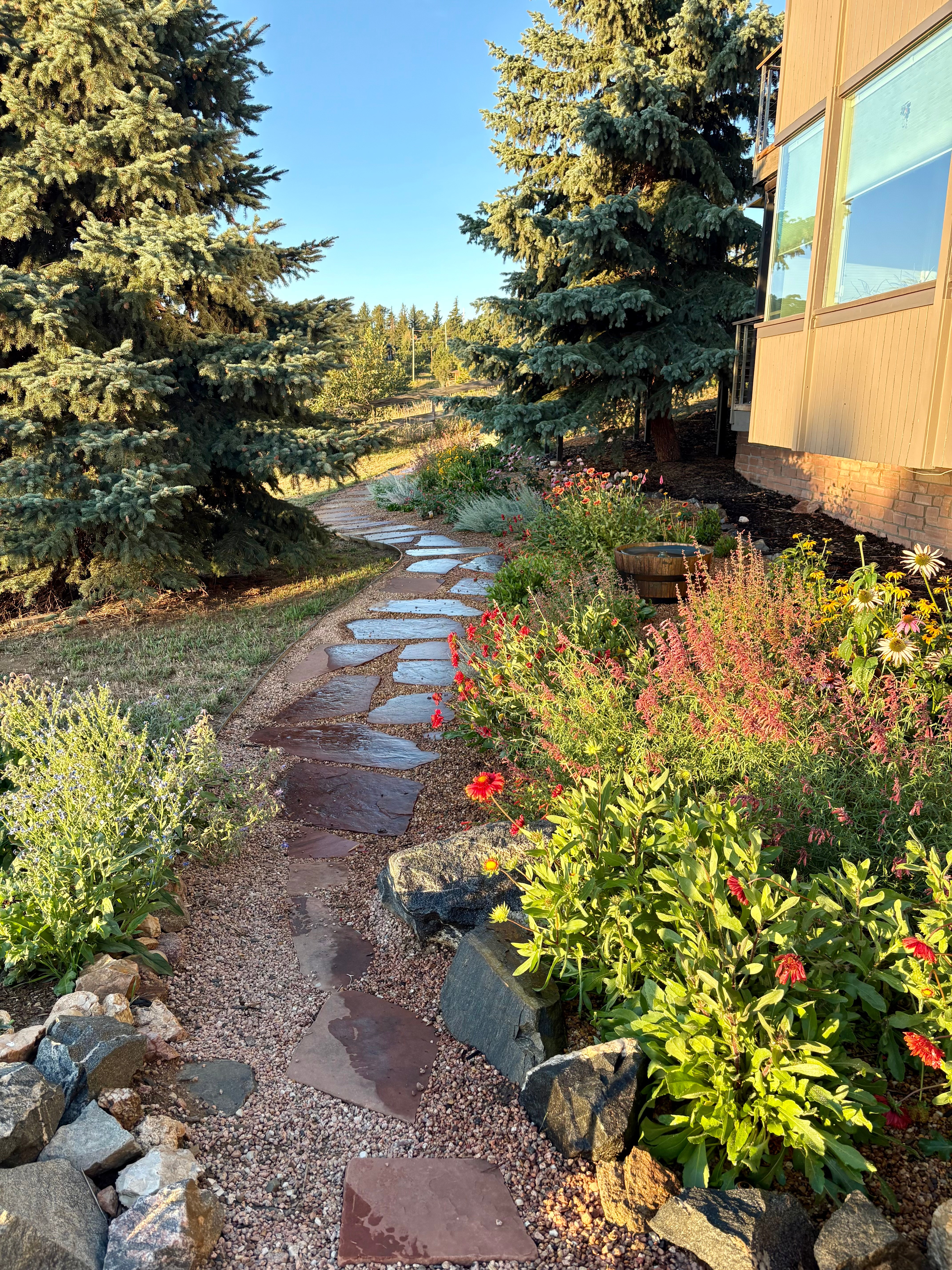 Stone pathway winding through a garden with colorful flowers and tall trees beside a building, under a clear blue sky.