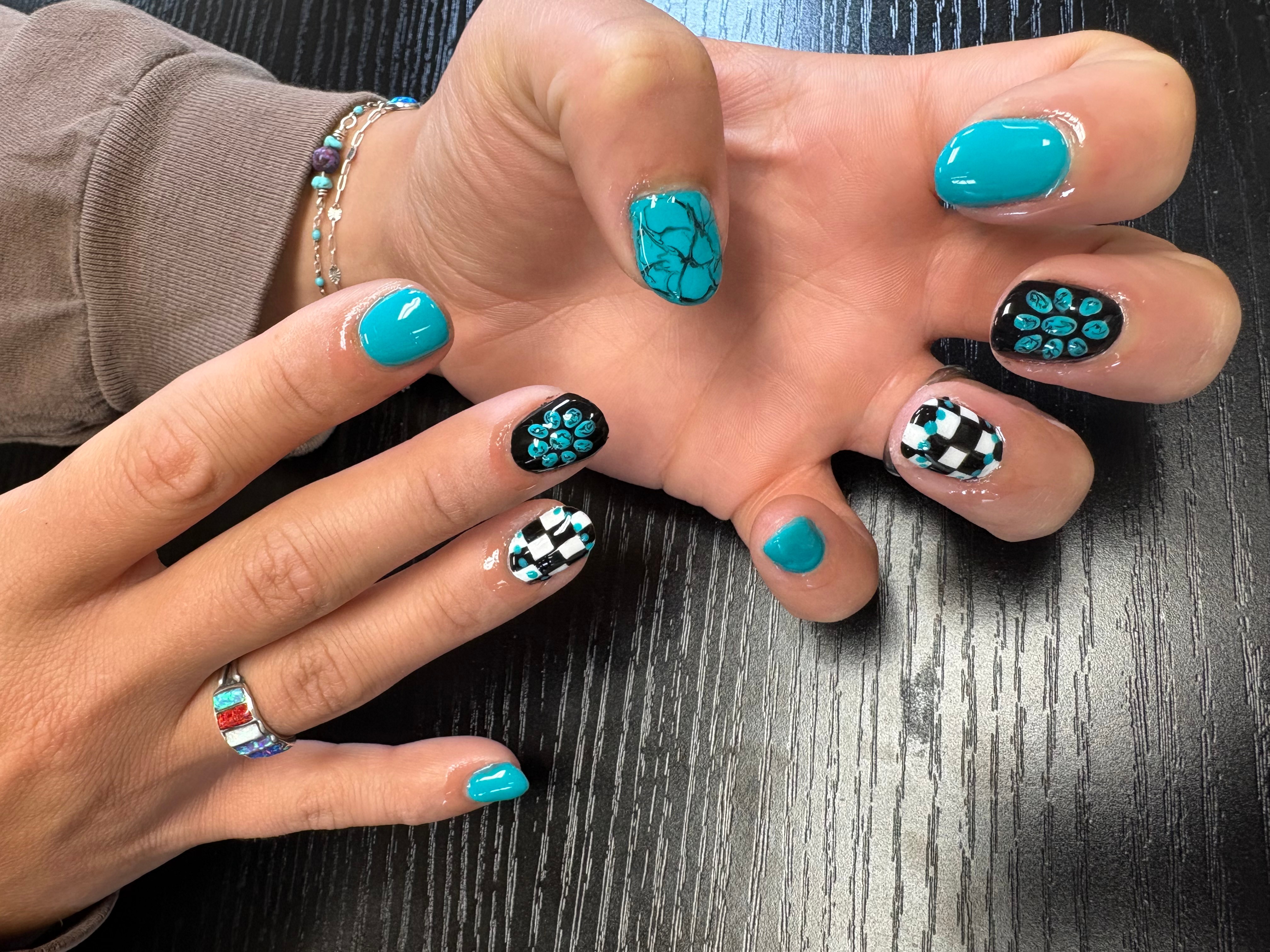 Two hands with turquoise, black, and checkered nail polish, resting on a dark wooden surface.