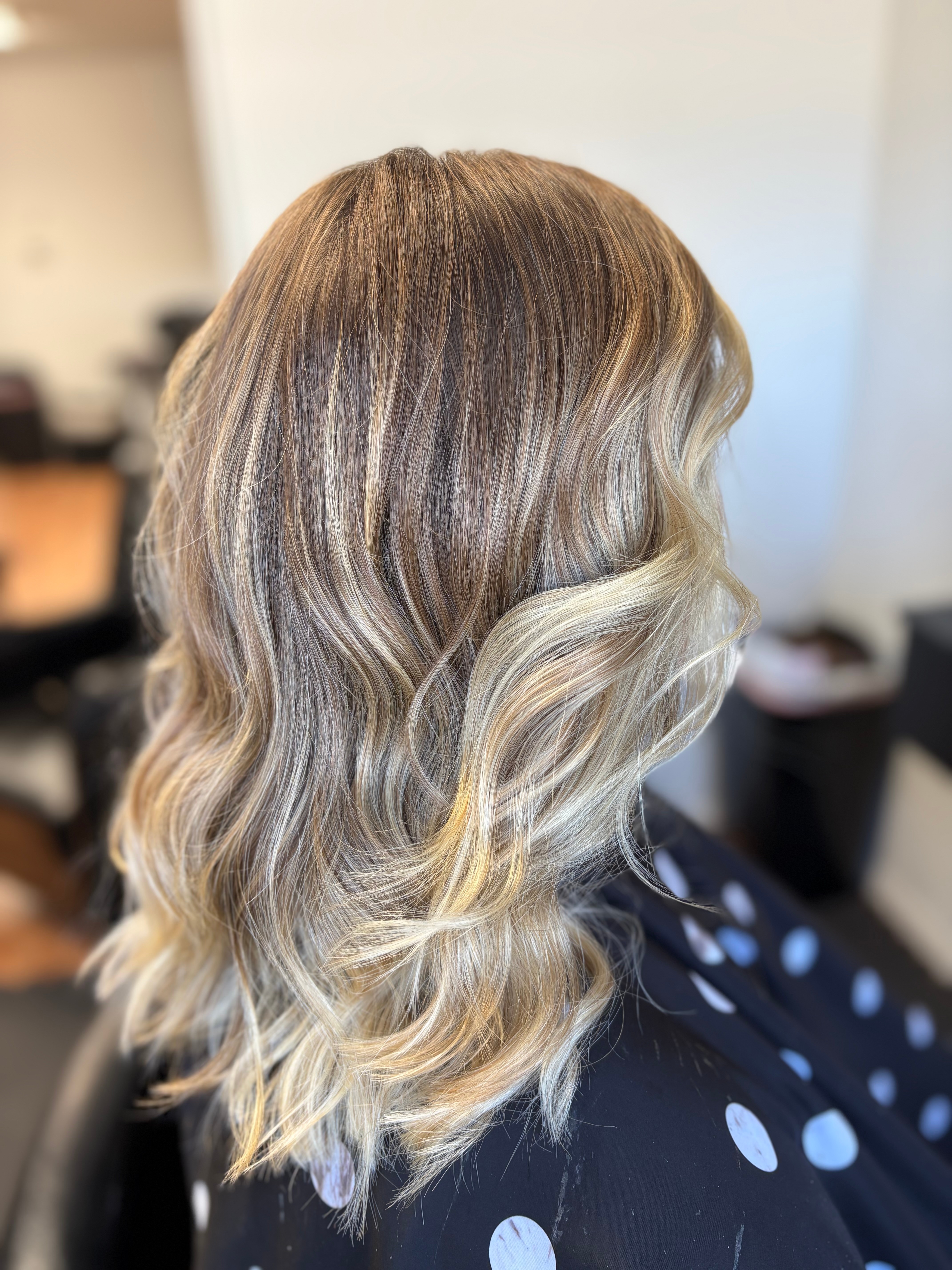 Side view of wavy, blonde hair with darker roots, styled in loose waves, against a blurred indoor background.
