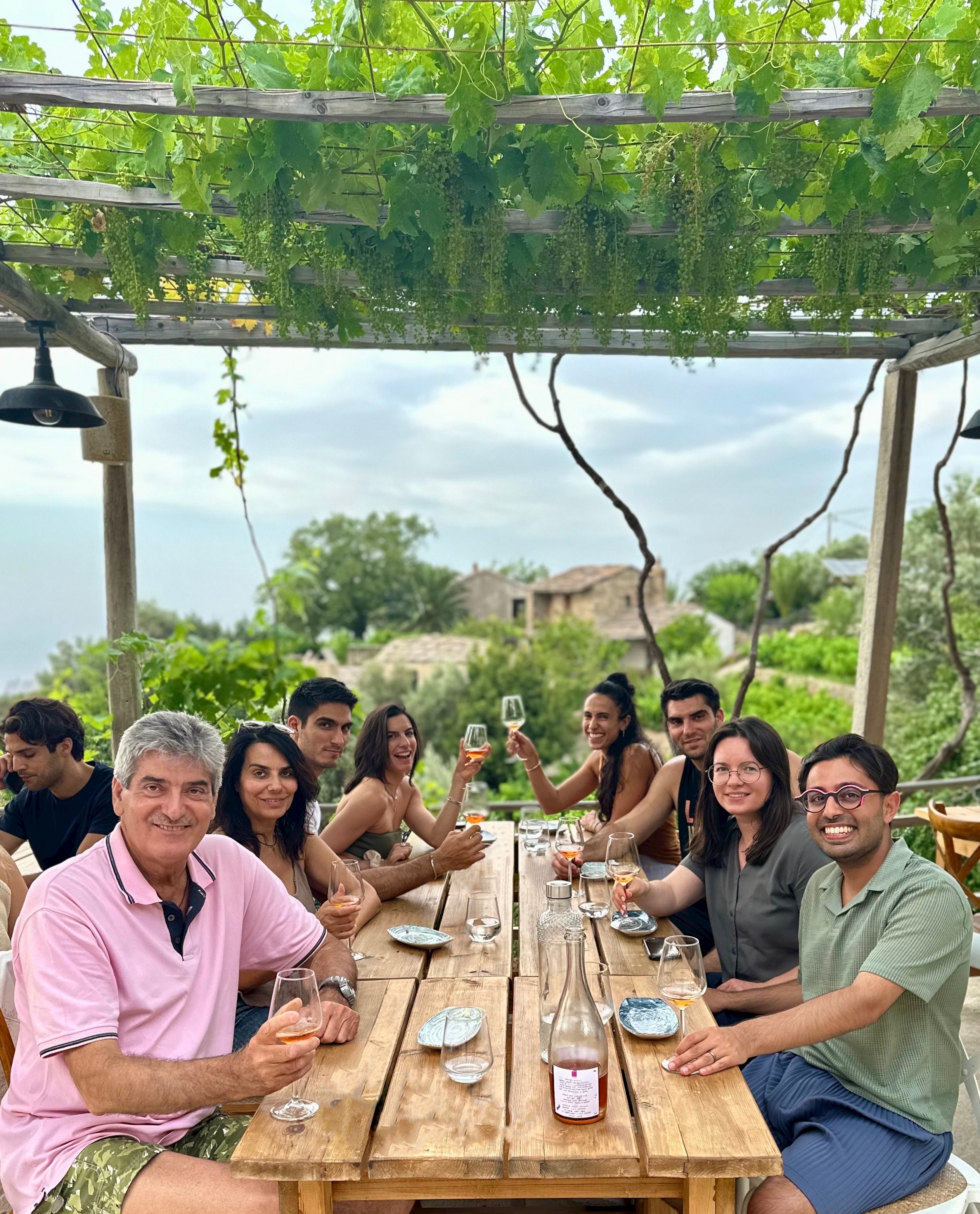 Group of people sitting at a long outdoor wooden table, smiling, with greenery and cloudy sky in background.