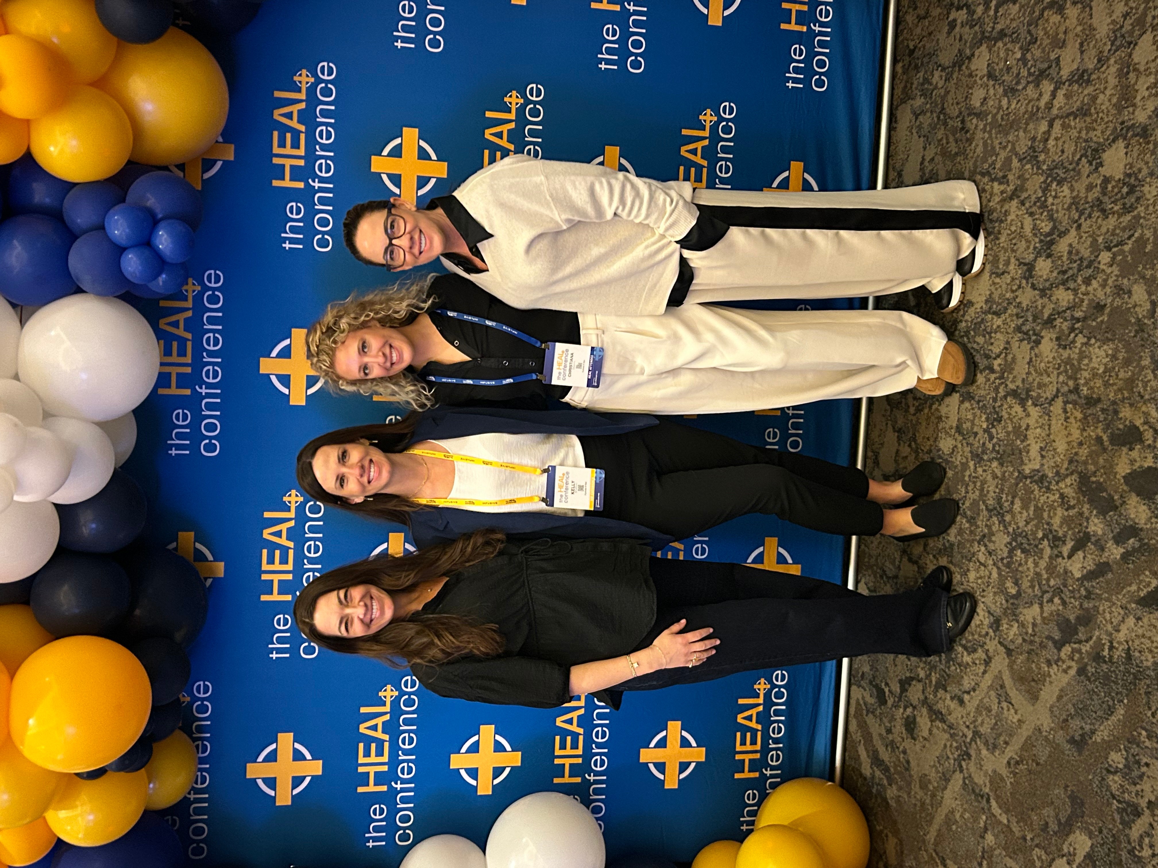 Three women standing in front of a blue backdrop with balloons, smiling at the conference.