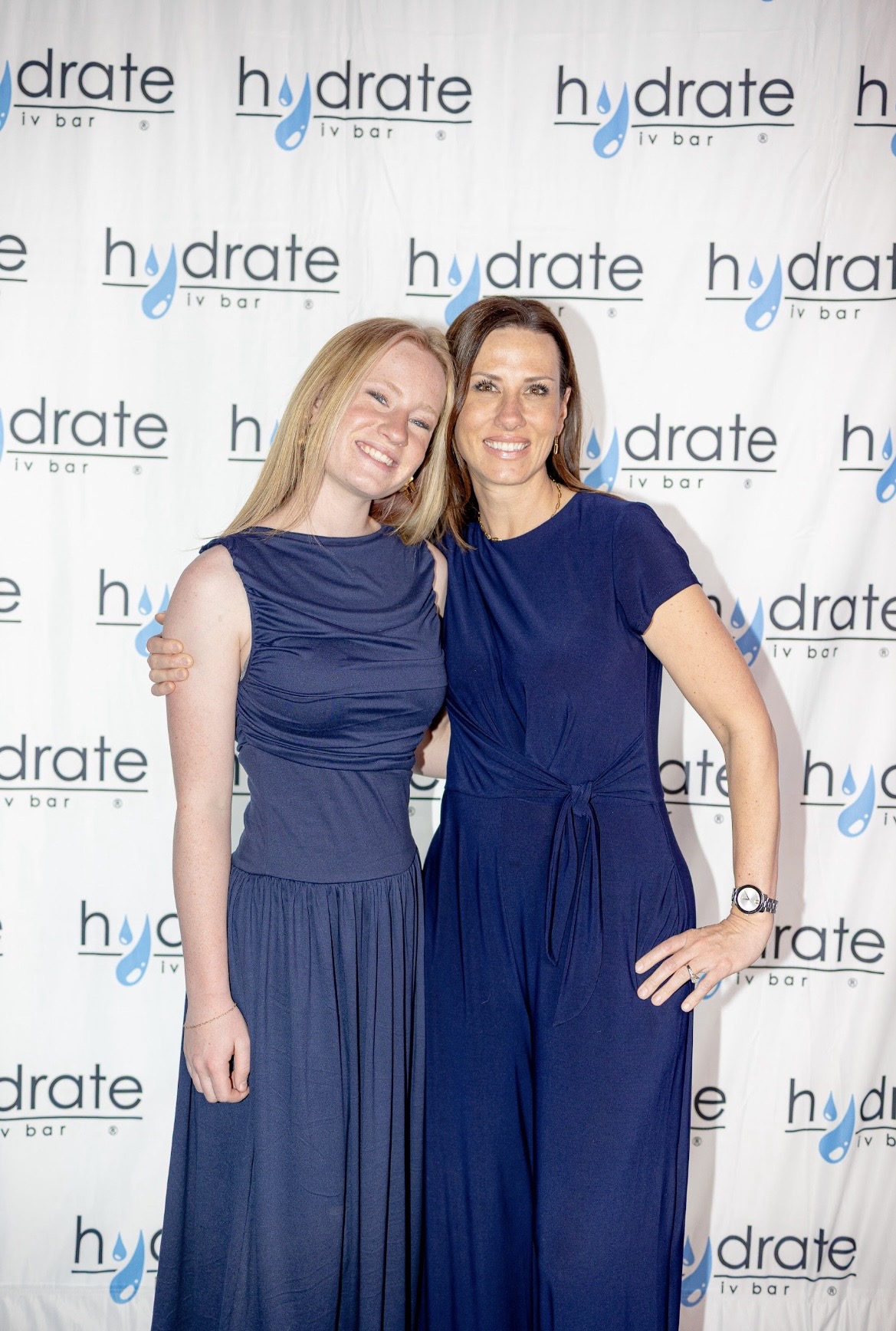 Two women in blue dresses standing together, smiling, in front of a backdrop with the word 'hydrate' repeated.