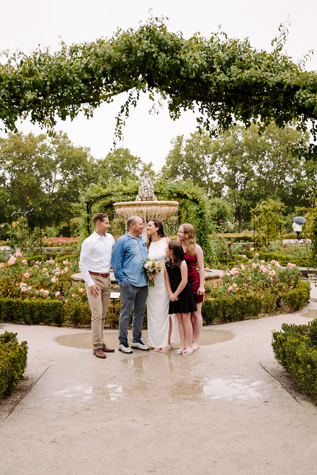 Group of six people standing in a garden with flowers and trees, under a large arching tree branch.