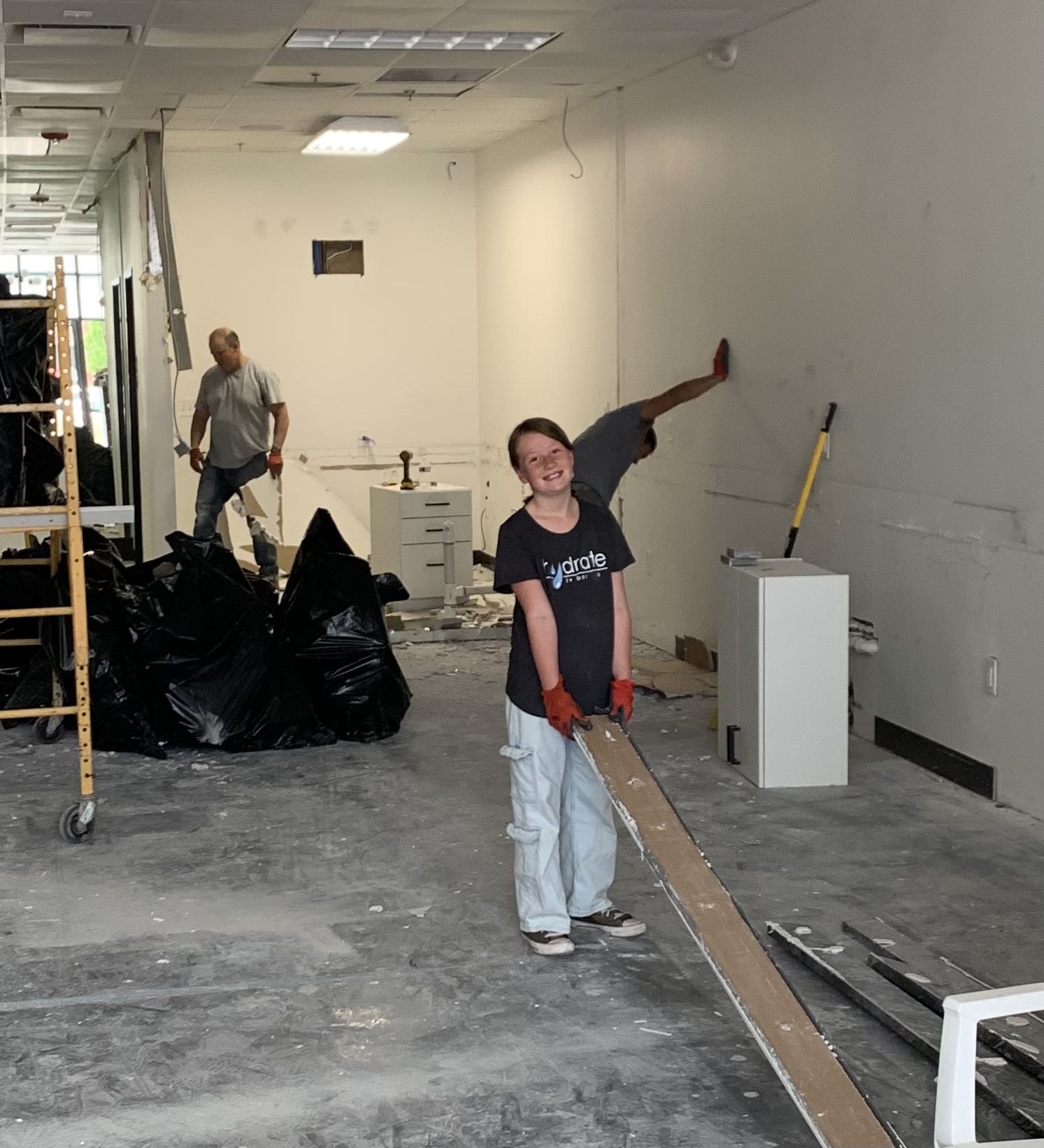 Young girl smiling, holding a long wooden plank, in an indoor construction or renovation space with two men working in the background.