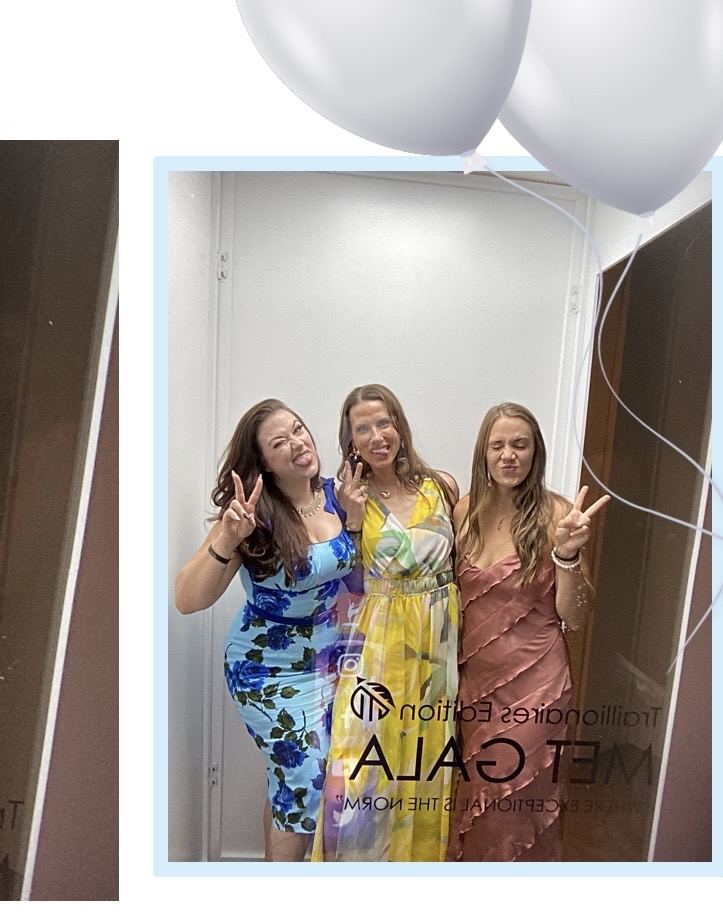 Three women smiling and posing inside an elevator, with balloons visible at the top, and a mirror reflecting their image.