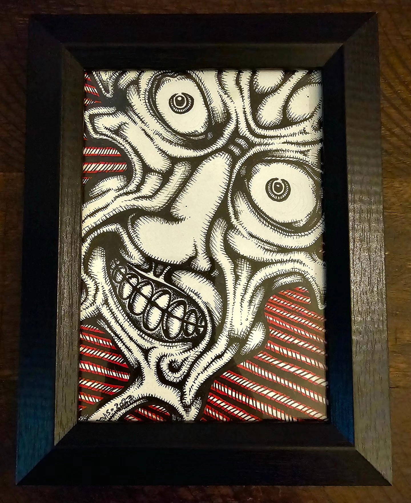 Black frame contains a detailed, exaggerated face with wide eyes and a large, toothy grin, against a red and white striped background.