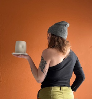Person with a tattoo on arm, wearing a gray beanie and black top, holding a candle on a plate against an orange background.