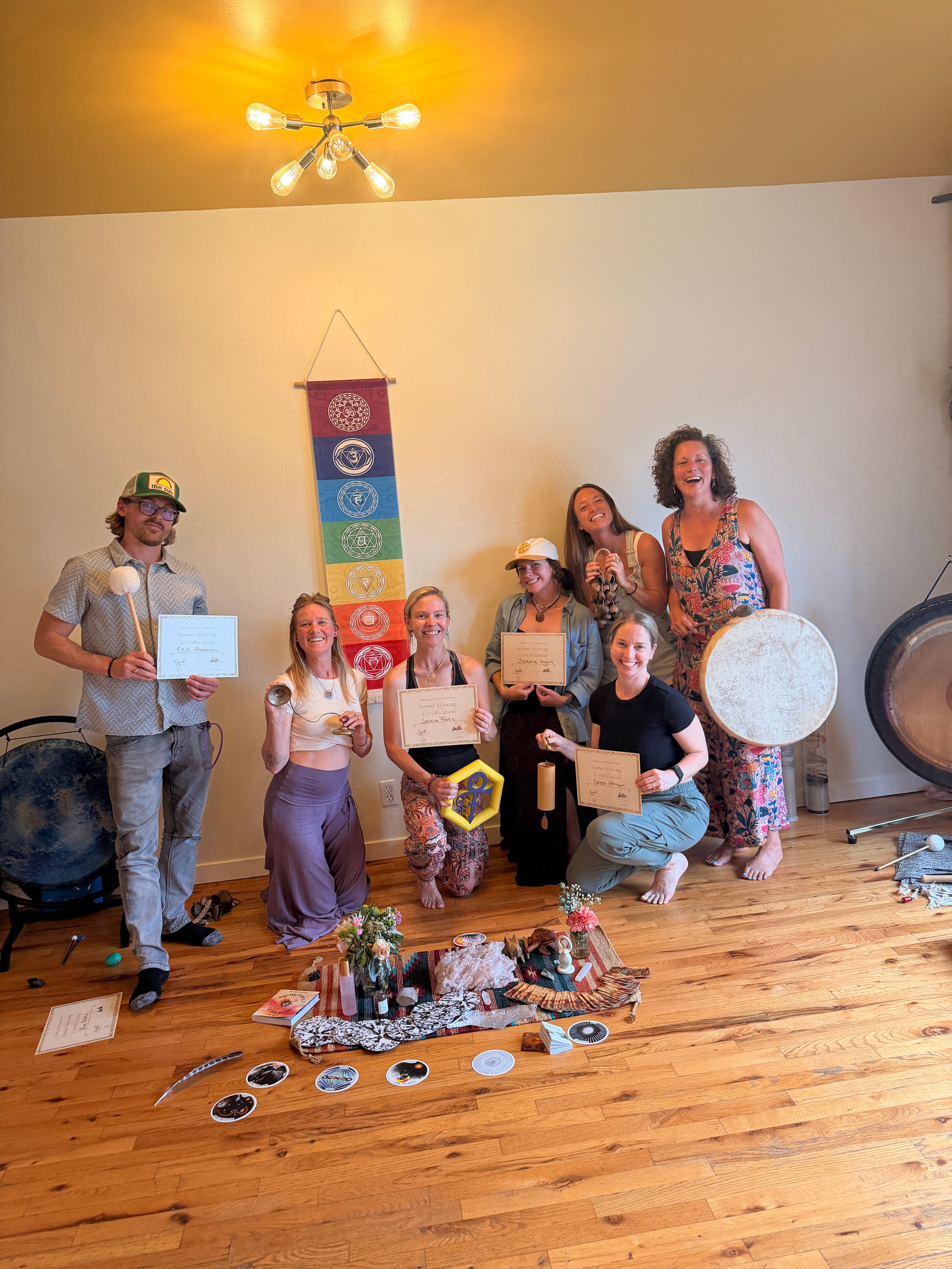Group of people standing and sitting on a wooden floor in a room with a yellow wall, colorful hanging decorations, and a chandelier.