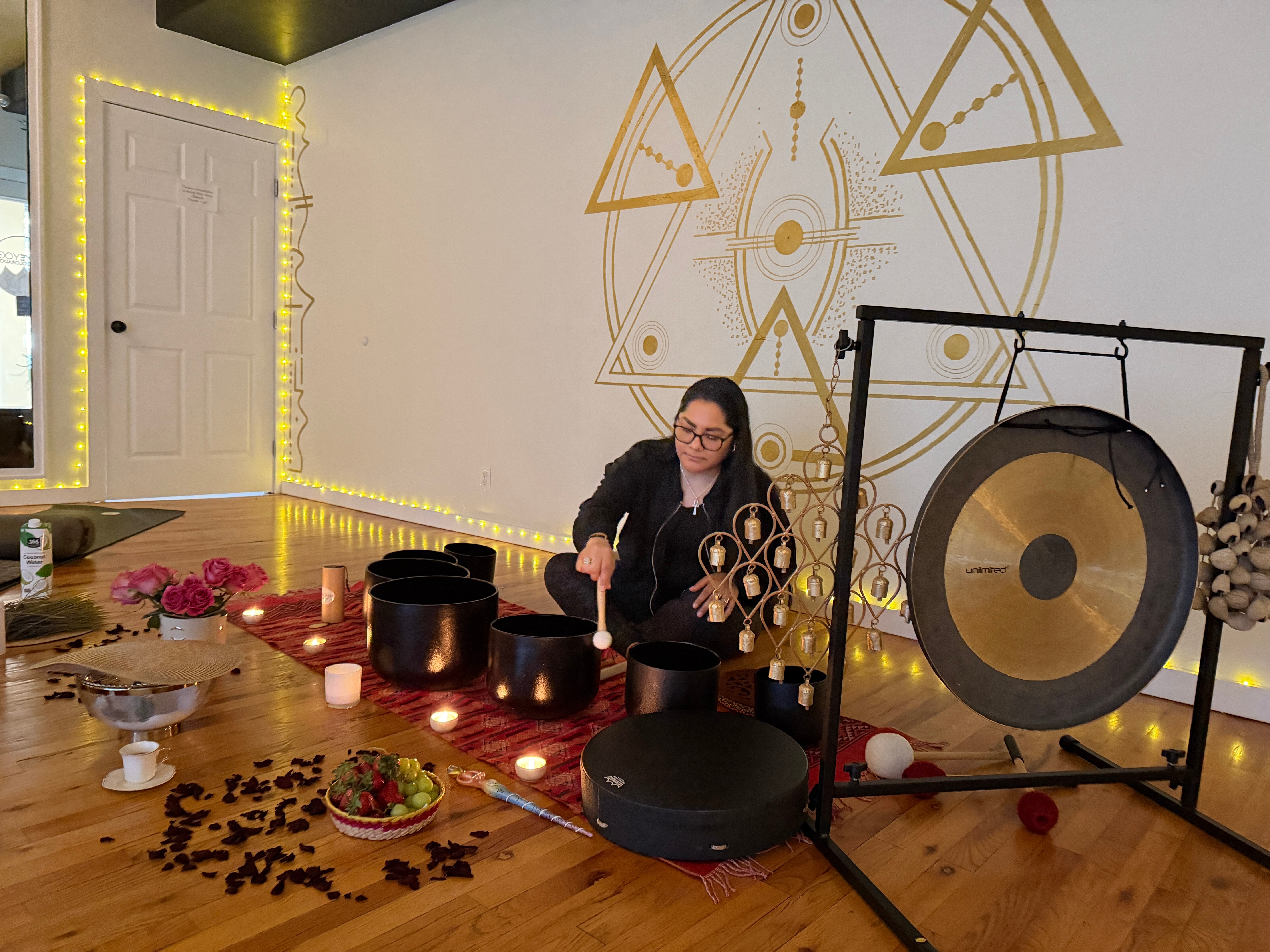 Person sitting on the floor with singing bowls, wall with geometric design, door, and string lights in the background.