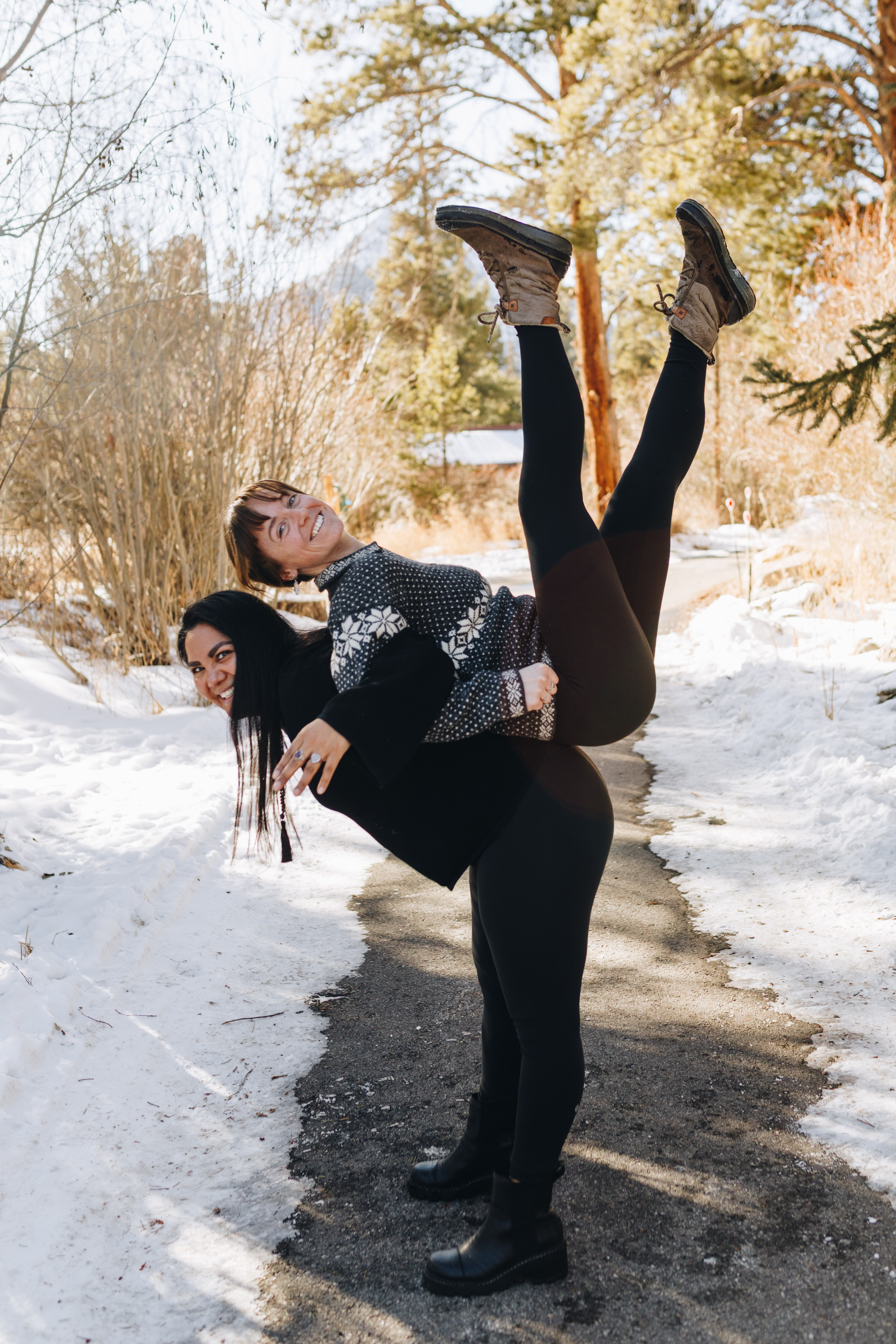 Two women outdoors on a snowy path, one lifting the other with legs in the air, trees in background.