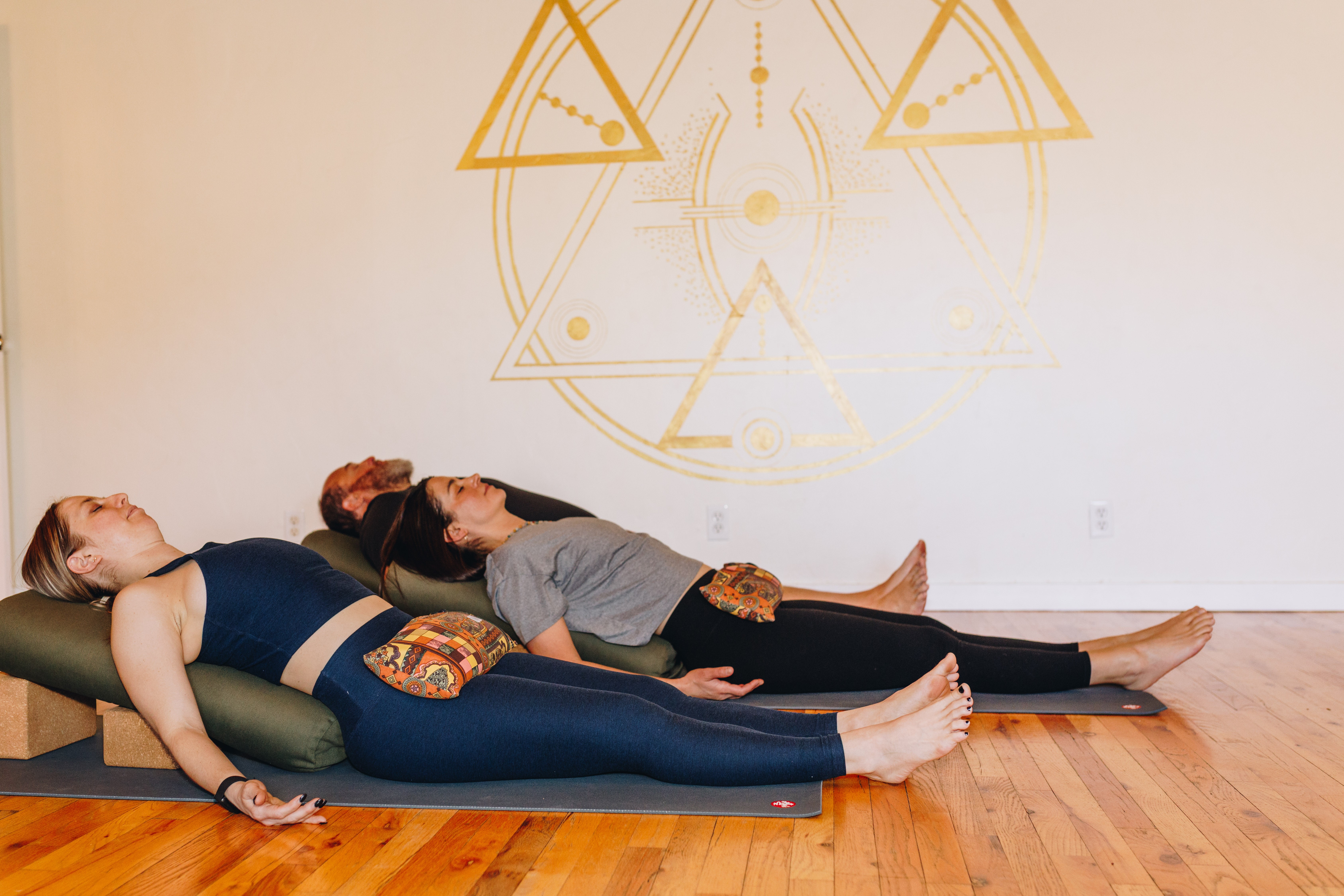 Three people lying on yoga mats in a studio, with a geometric design on the wall behind them.