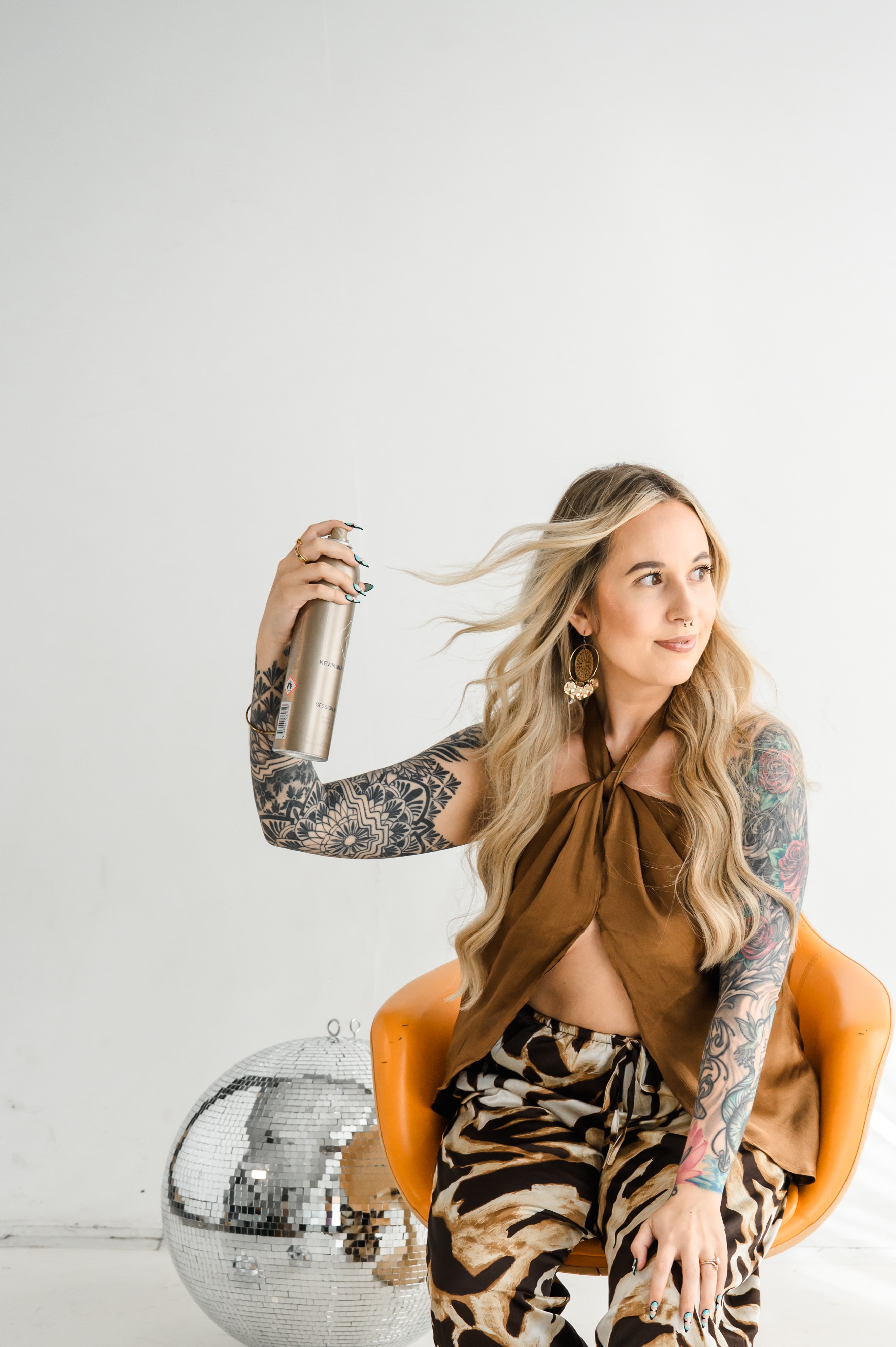 Woman with long hair and tattoos holding a spray bottle, sitting on an orange chair against a plain white background.