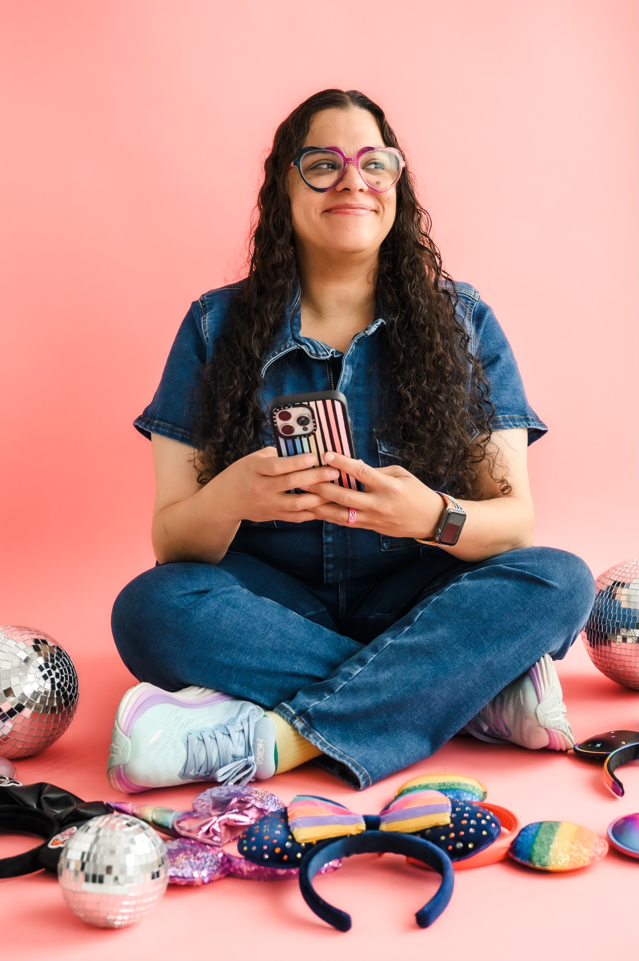 Person sitting cross-legged on pink background, holding a phone with an American flag case, smiling, wearing glasses and denim outfit.