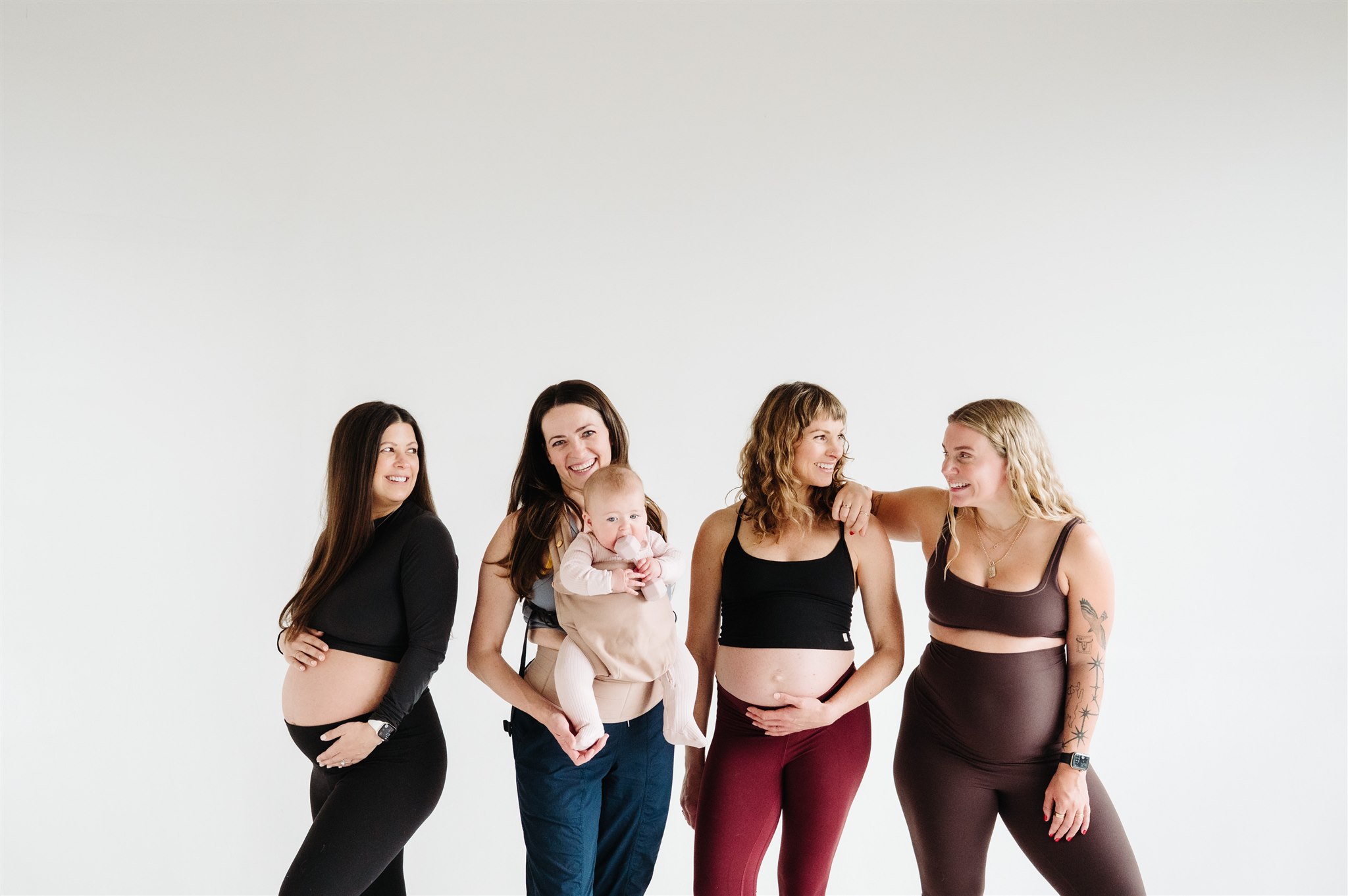 Four women, one holding a baby, standing against a plain white background, engaged in conversation, smiling, and looking at each other.