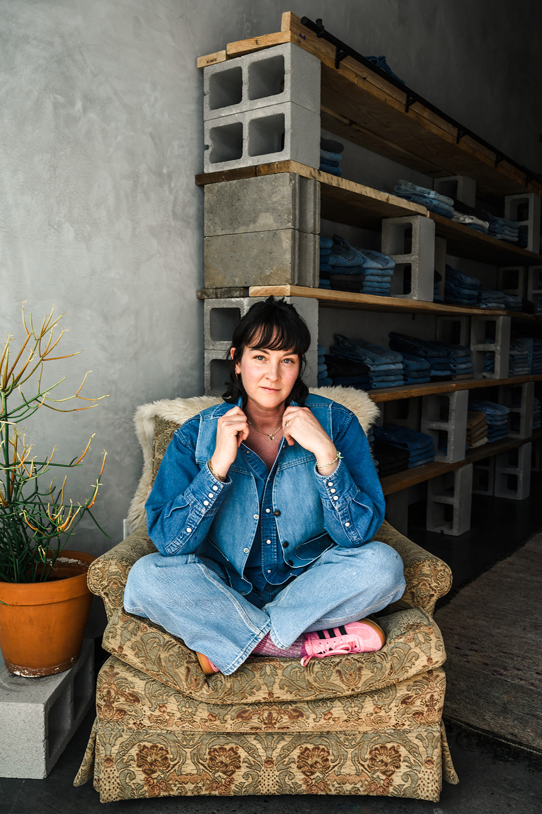 Young woman sitting cross-legged on a patterned armchair, wearing a denim jacket and jeans, in a room with shelves and a potted plant.