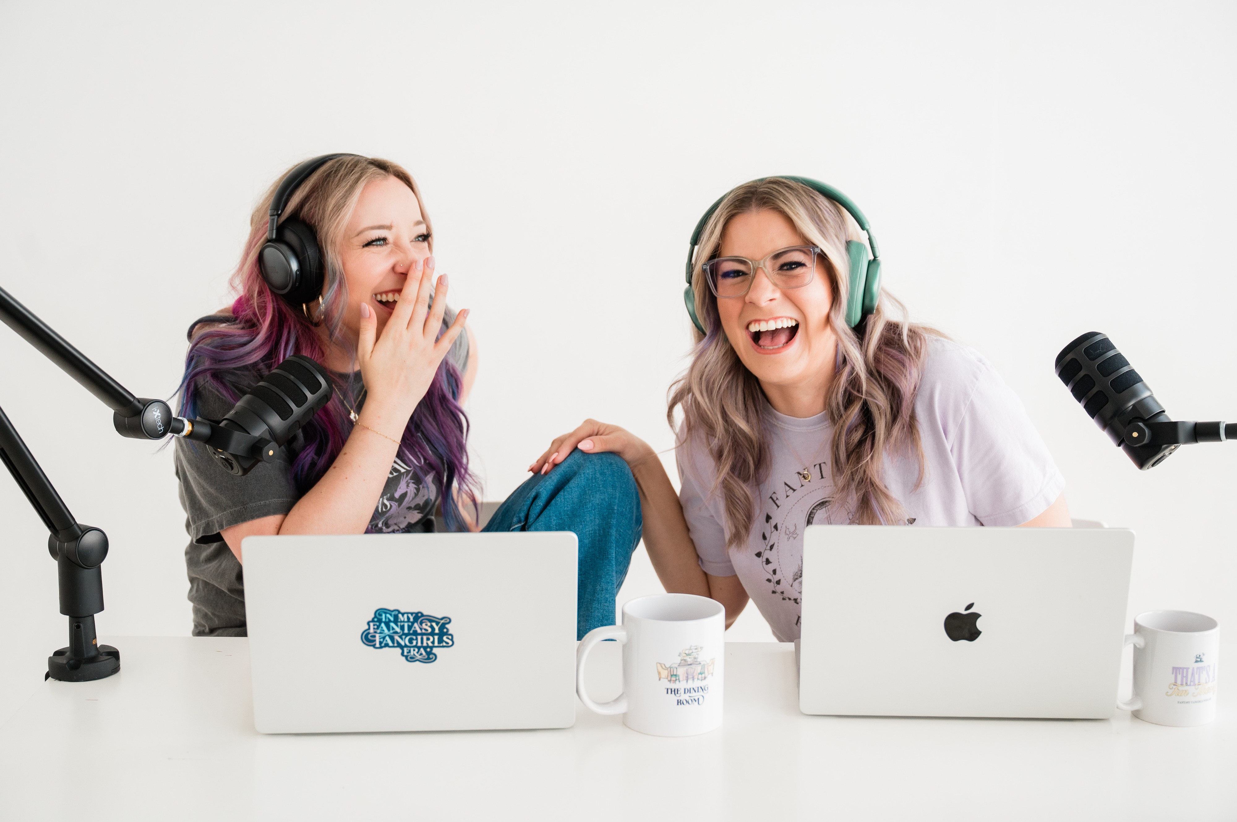 Two women with headphones laughing and talking in front of laptops and microphones, white background.