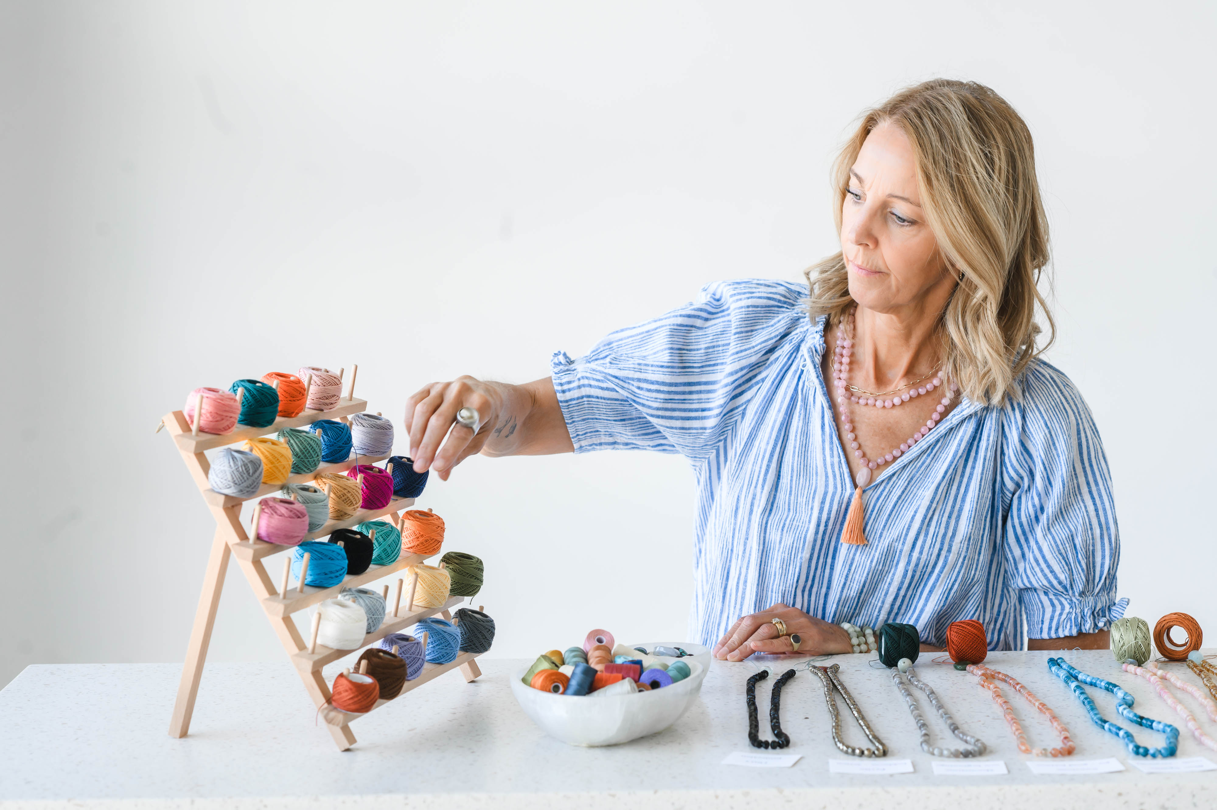 Woman with blonde hair in a blue striped shirt arranges colorful yarn on a wooden stand, with bowls and jewelry on table.