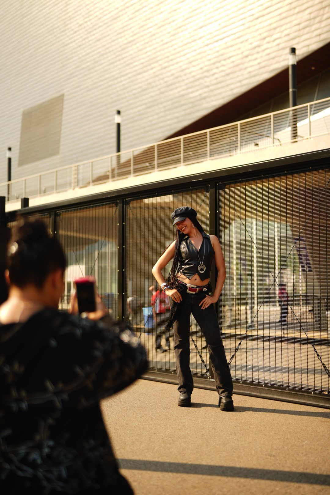 Person standing on a balcony with a railing, holding a phone, with a building and window behind.