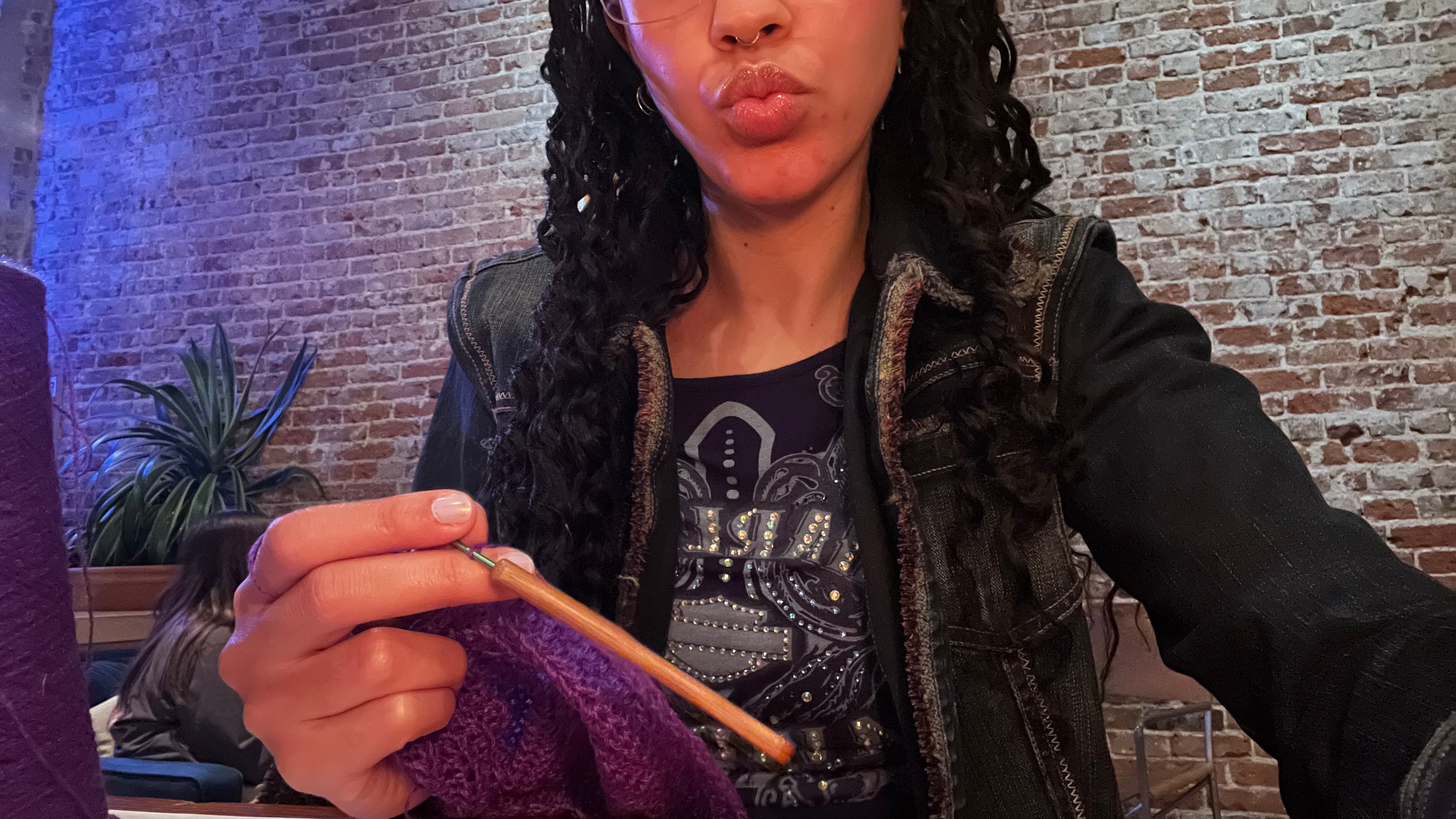 Person with curly hair making a kiss face, holding a purple object, in a brick-walled setting.