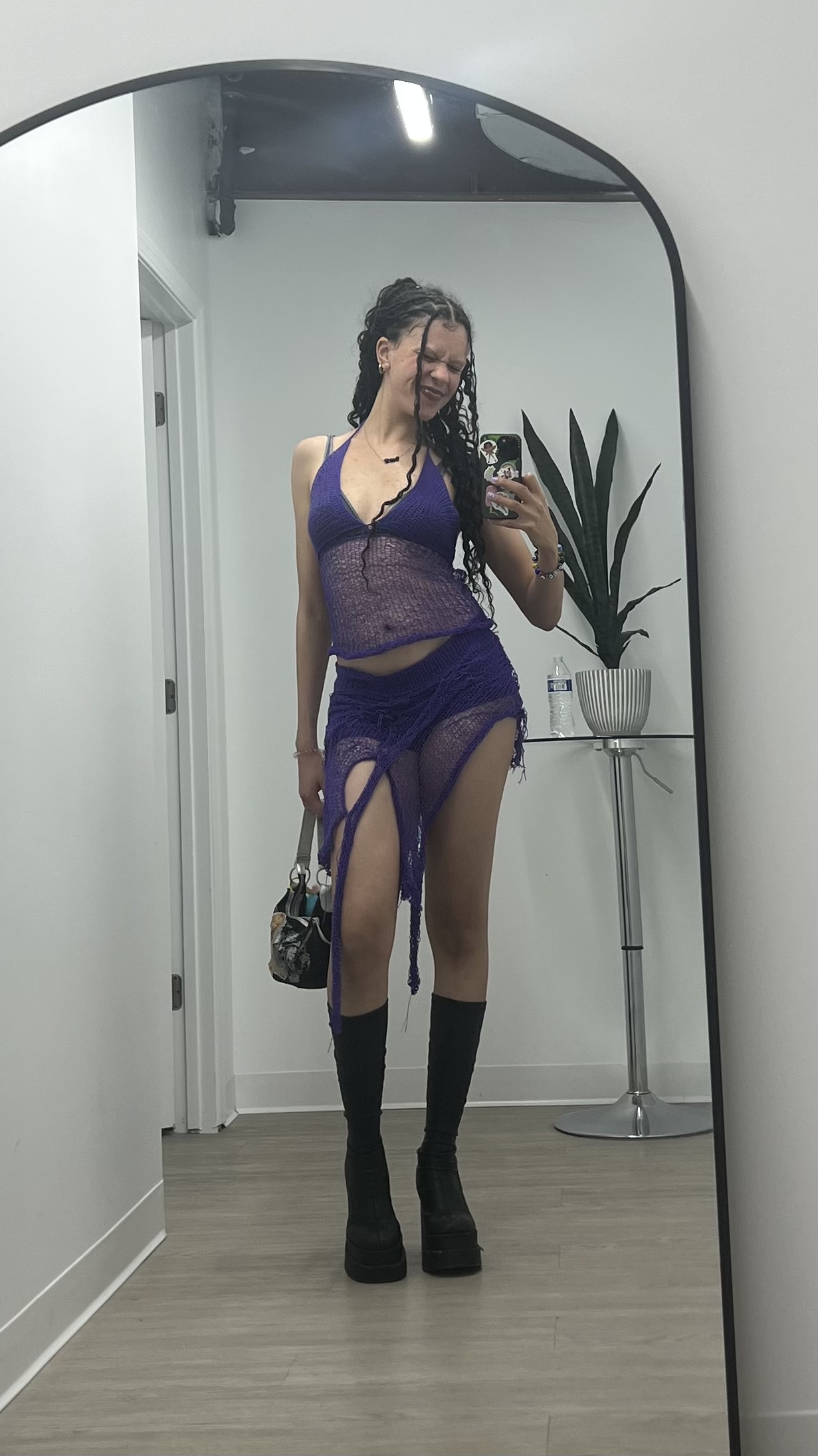 Person taking a mirror selfie in a room with a plant, wearing a purple outfit and black boots.