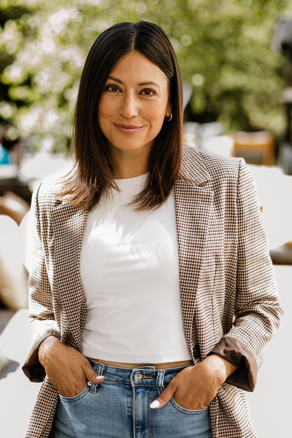 Woman with dark hair wearing a checkered blazer and white t-shirt outdoors, smiling, with trees in background.