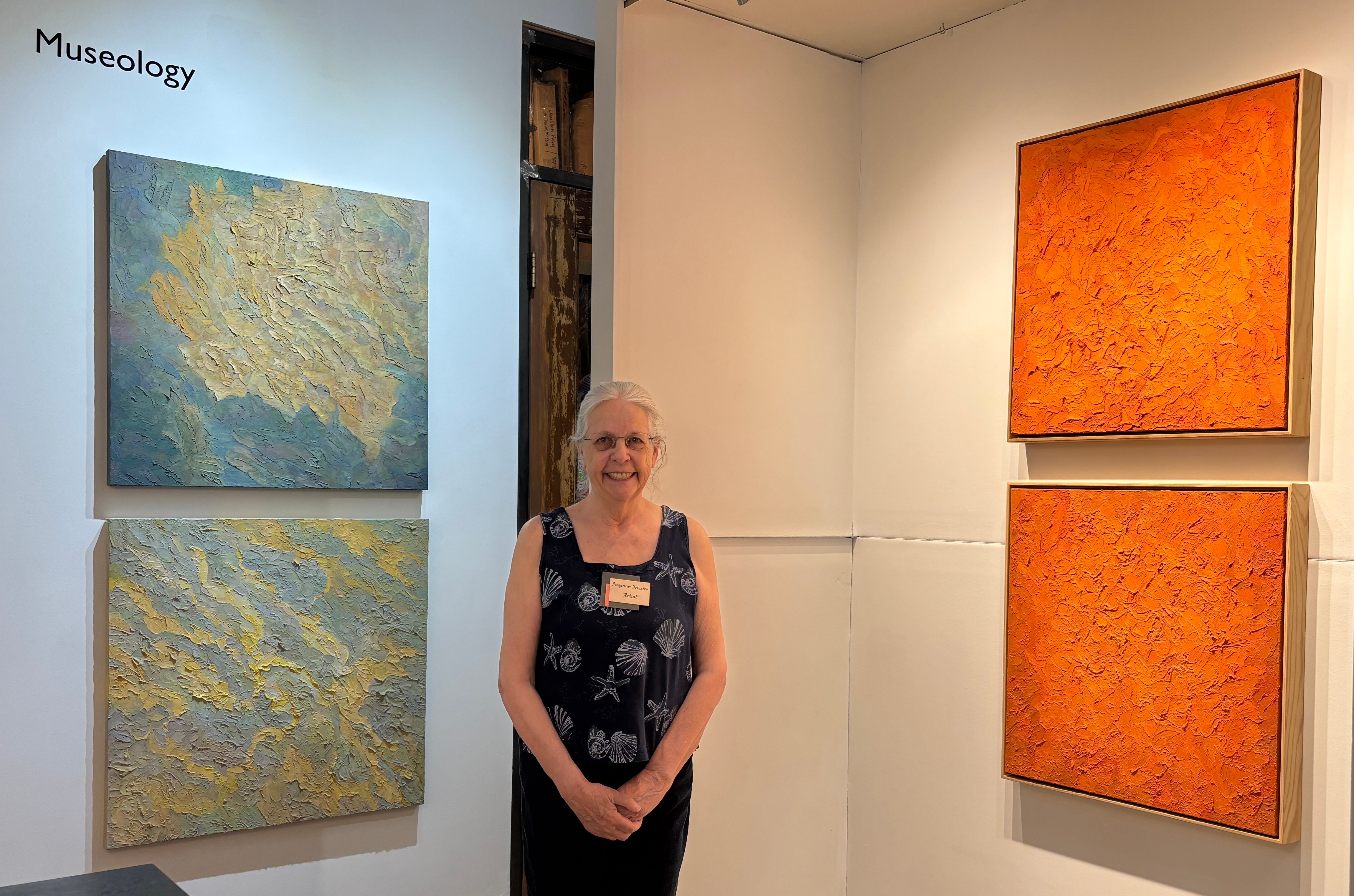 Woman standing in art gallery with four abstract paintings on white walls, two on each side, one set with blue and yellow, the other orange.