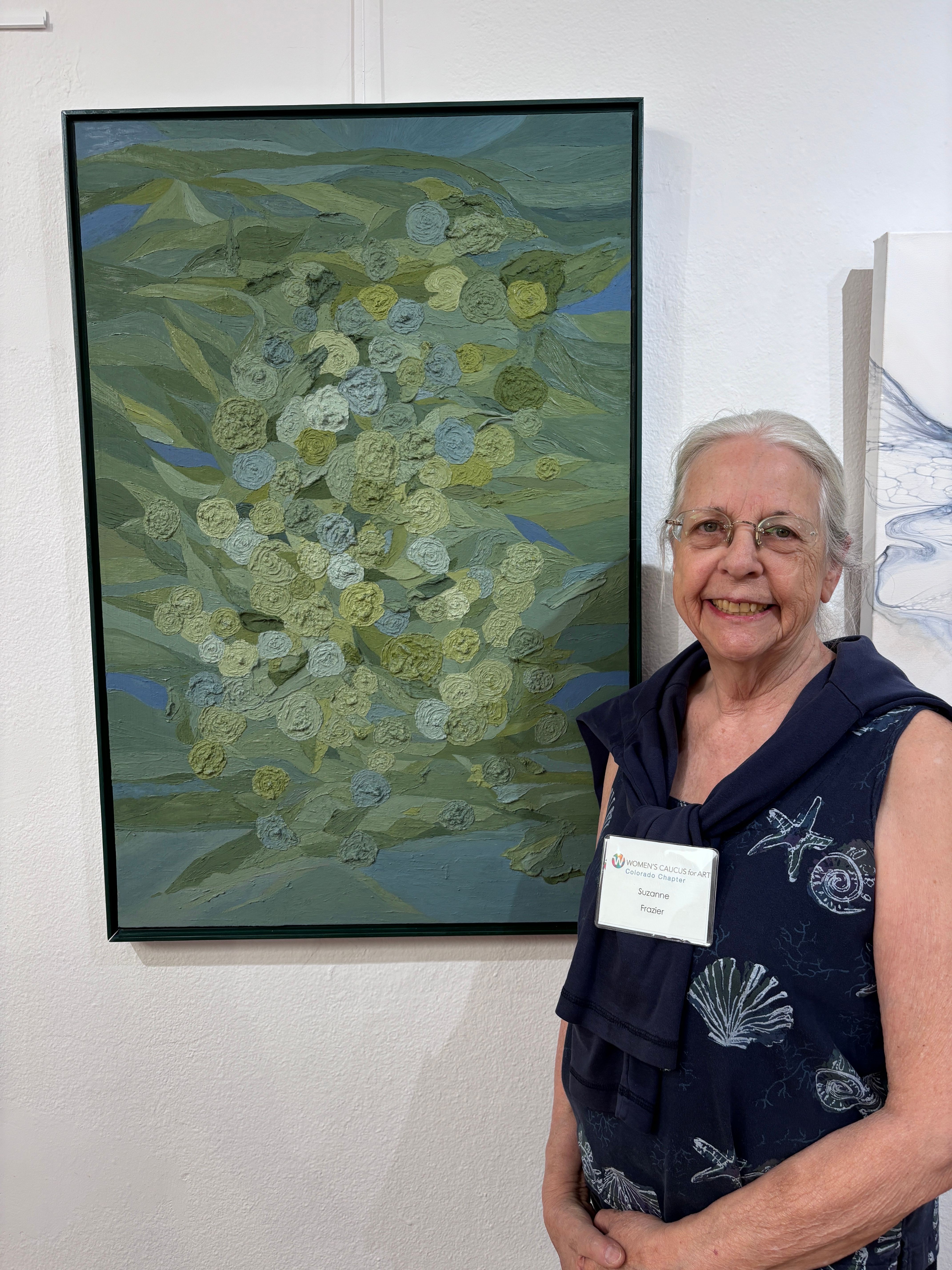 Elderly woman with glasses and short gray hair standing next to a large framed painting of green and white flowers, smiling.