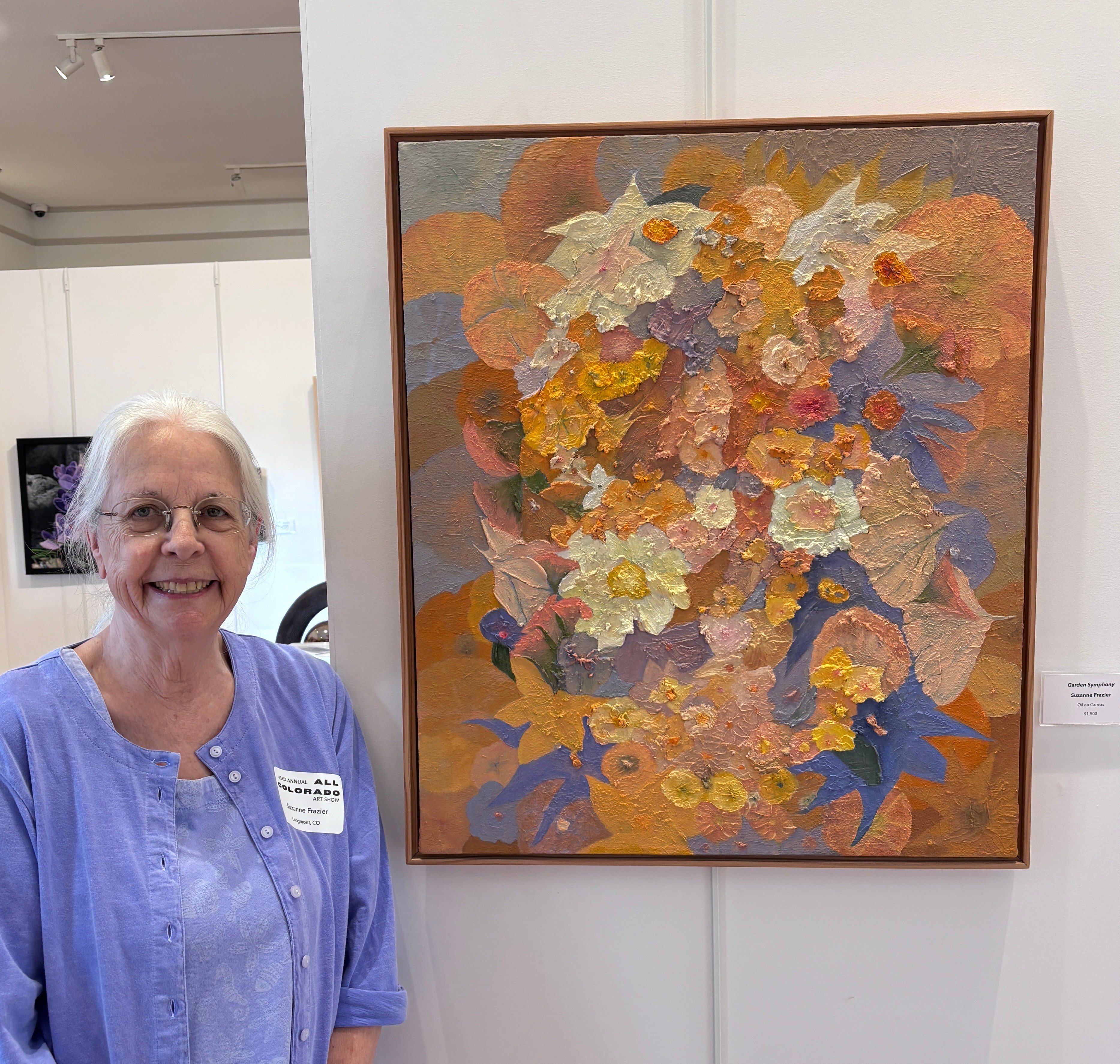 Elderly woman with glasses and a blue top standing next to a colorful floral painting on a white wall.