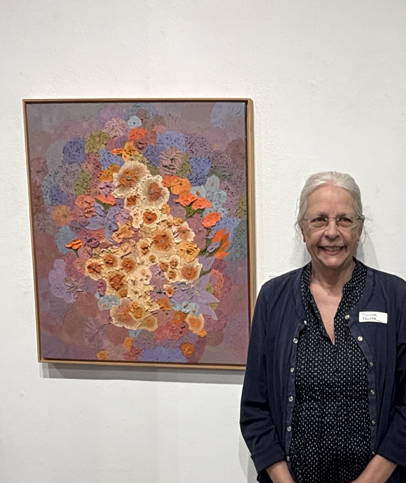 Older woman with white hair and glasses smiling, standing next to a colorful floral painting on a white wall.