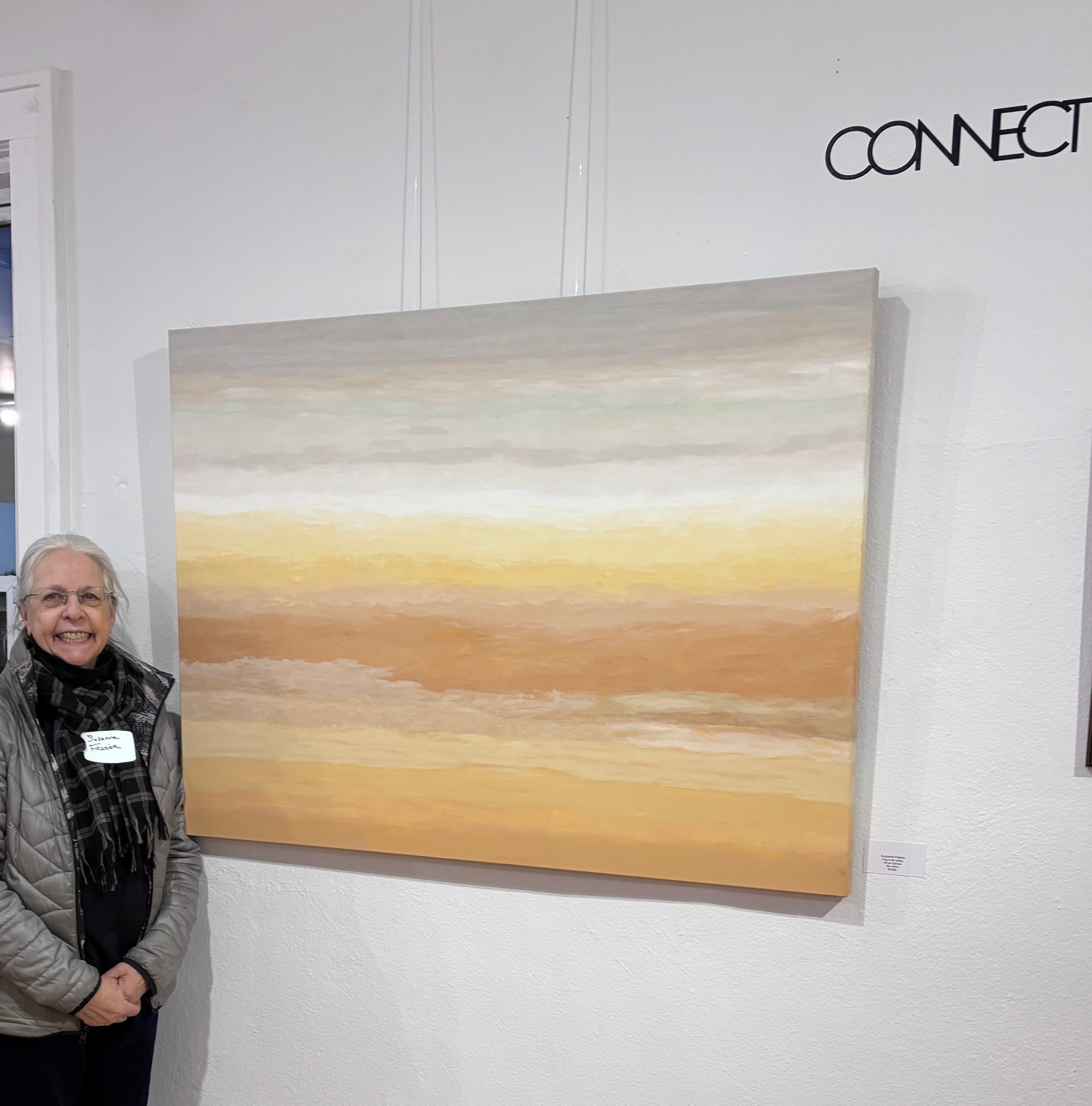 Person with glasses and a scarf standing next to a large abstract painting with horizontal bands of soft colors, on a white wall.