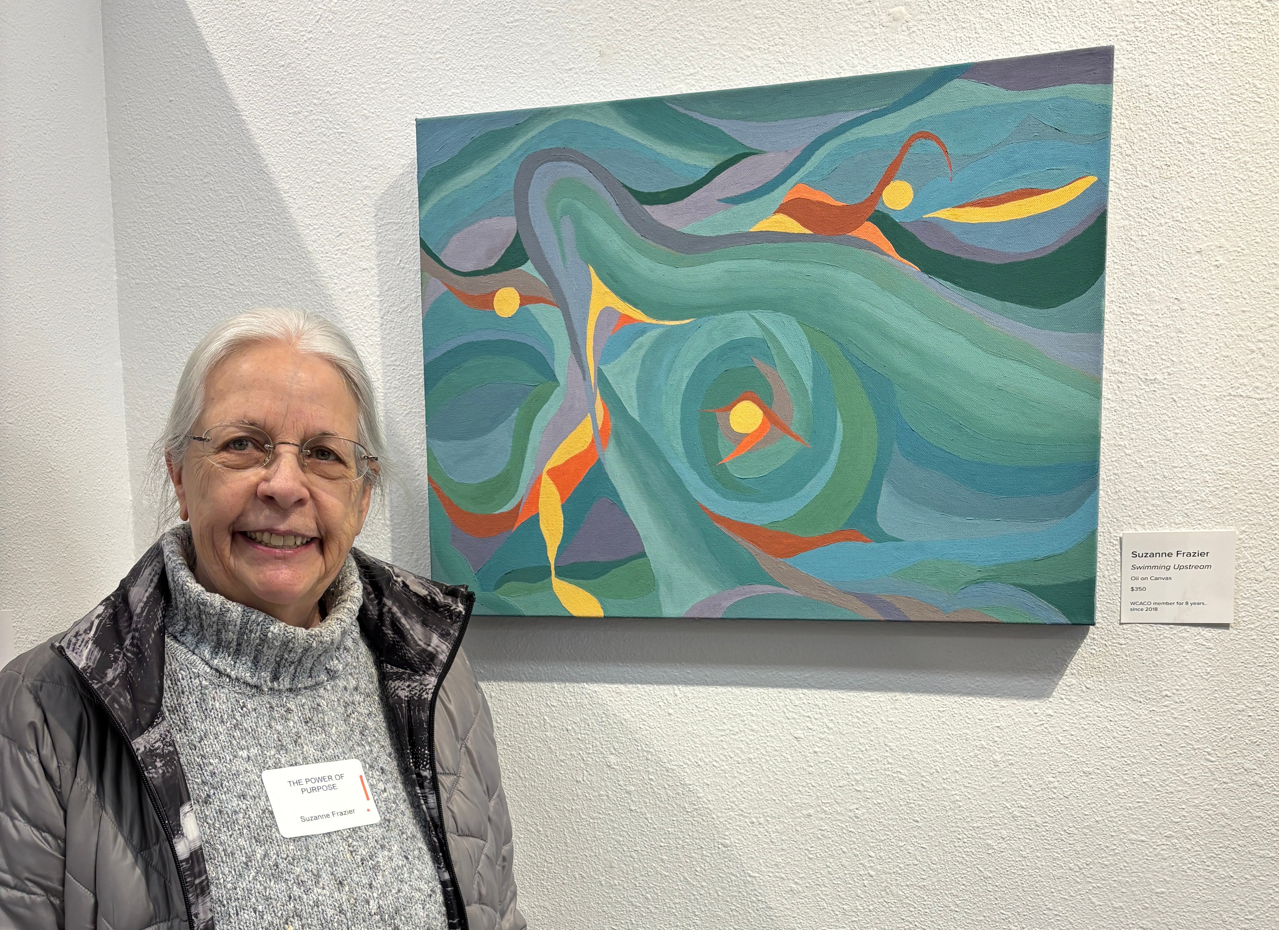 Older woman with glasses smiling in front of an abstract colorful painting on a white wall.