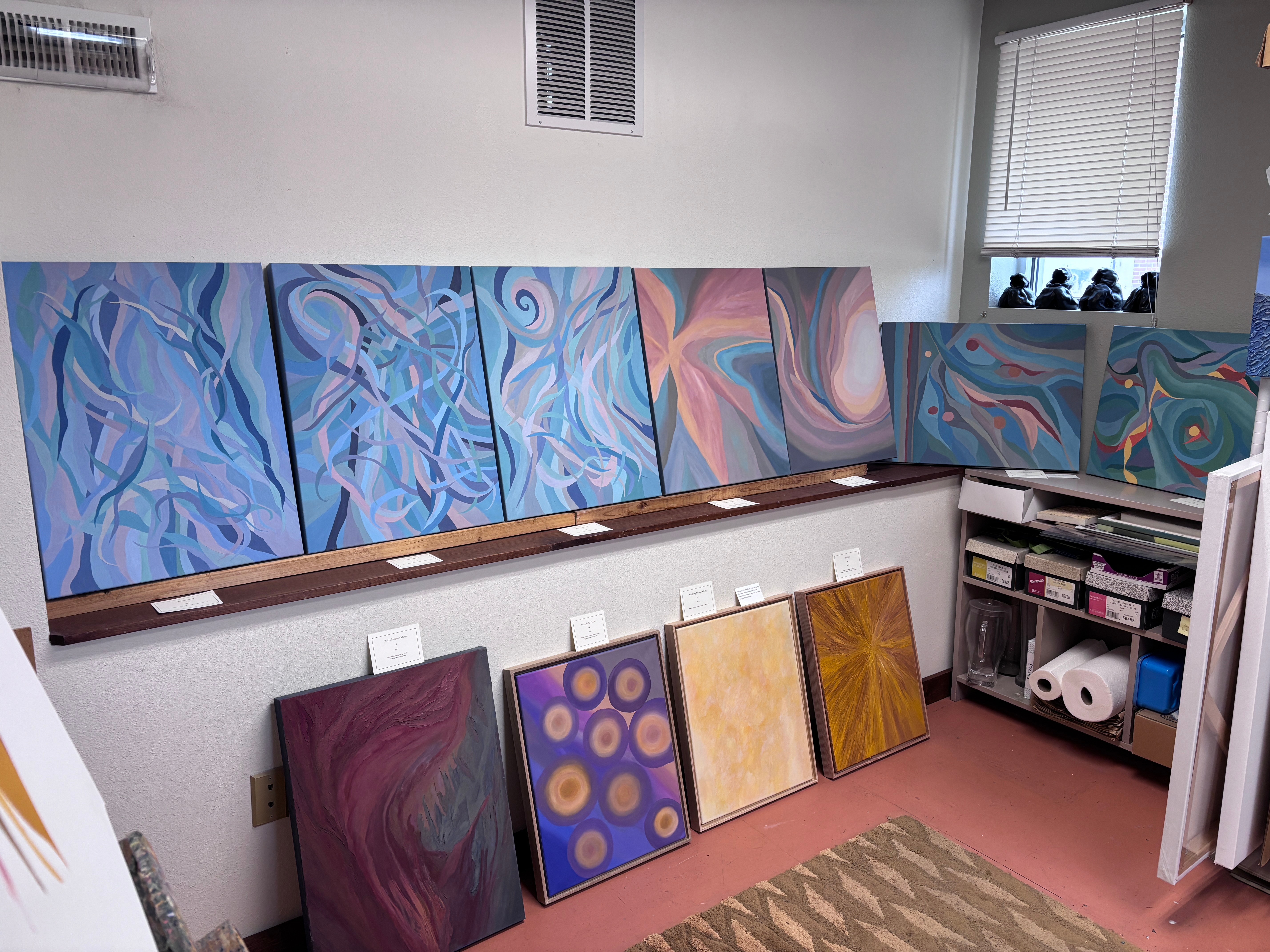 Multiple abstract paintings with swirling patterns and vibrant colors are displayed on a wall in an art studio.