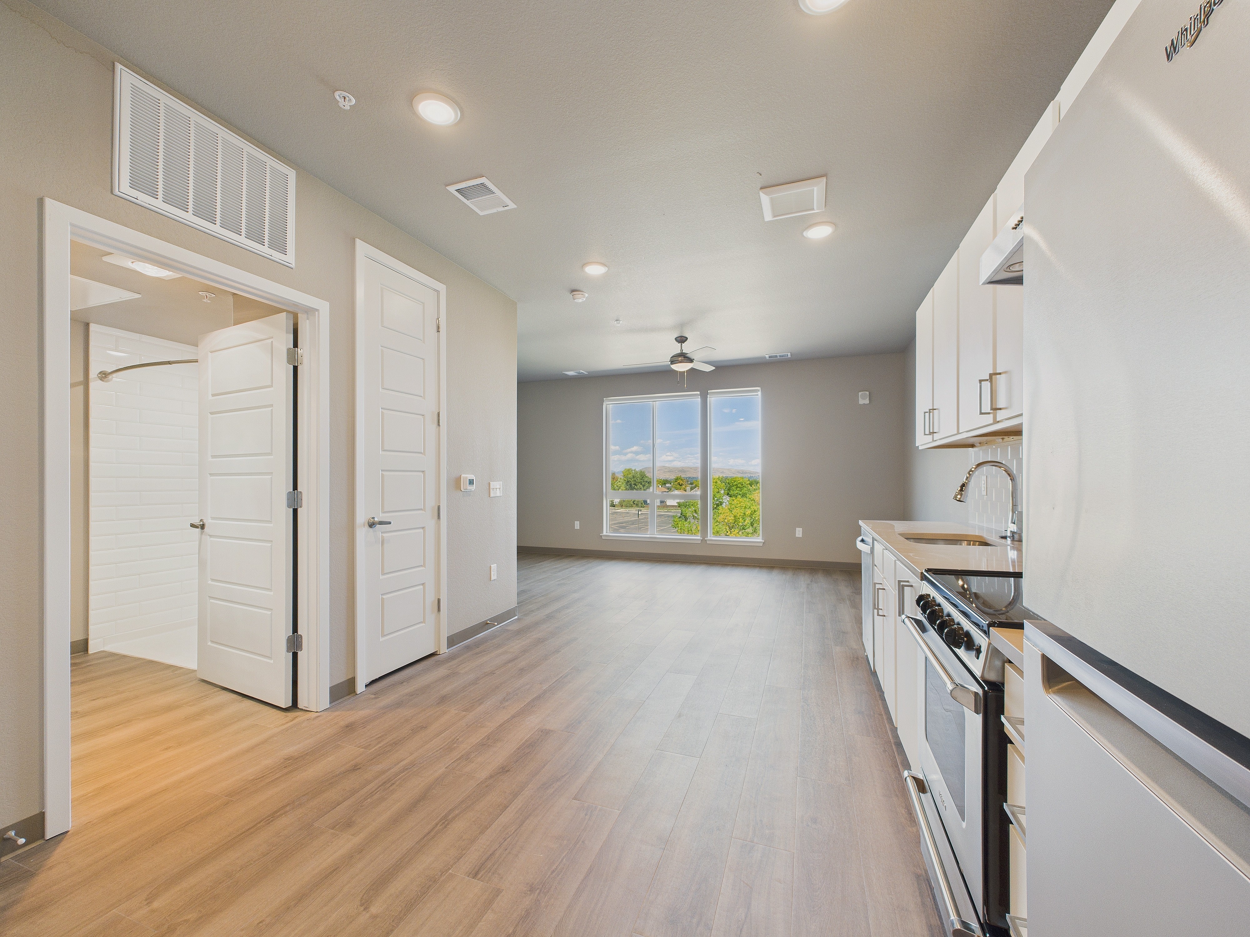 Empty room with kitchen area, large window, ceiling fan, light wood flooring, white walls, and cabinets.