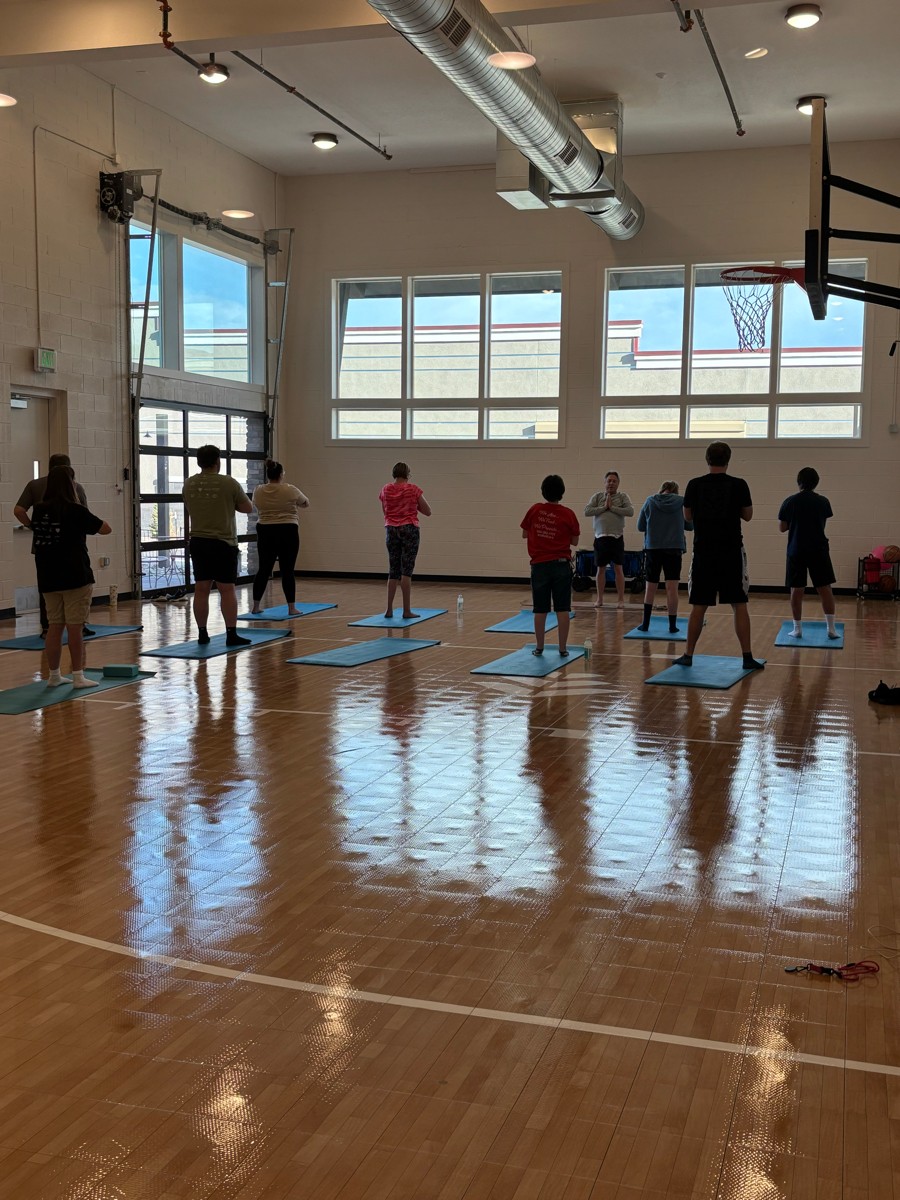 People practicing yoga or stretching on mats in a gymnasium with large windows and wooden floor.