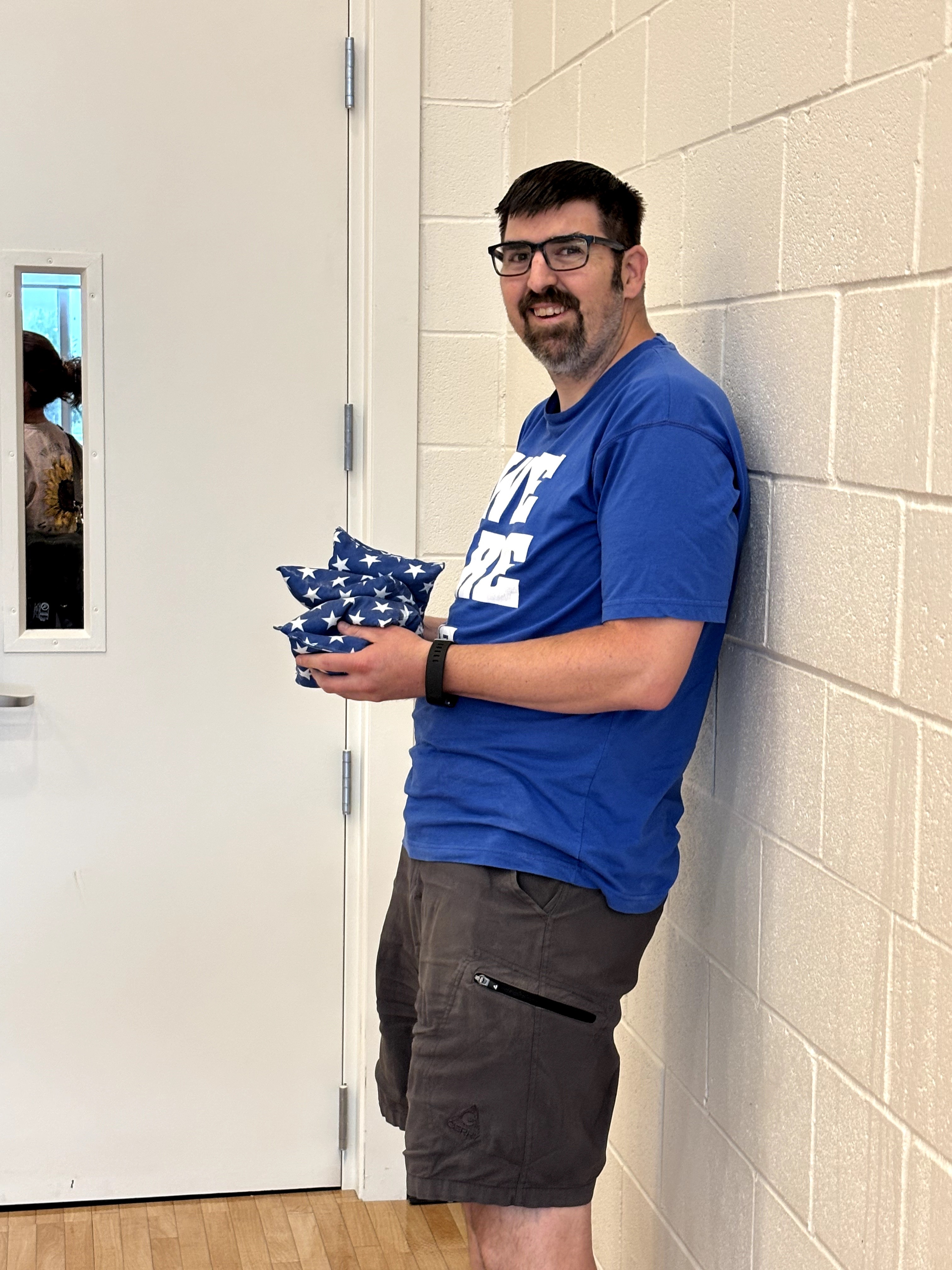 Man with glasses in blue shirt and shorts leaning against a wall, holding a wrapped gift, smiling.