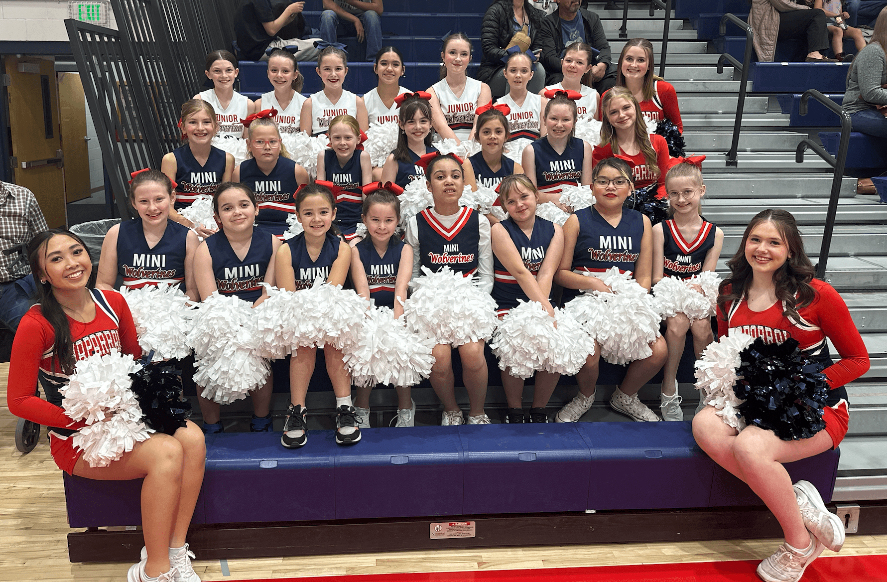 Group of young cheerleaders in uniforms holding pom-poms, seated and standing on bleachers in a gymnasium.