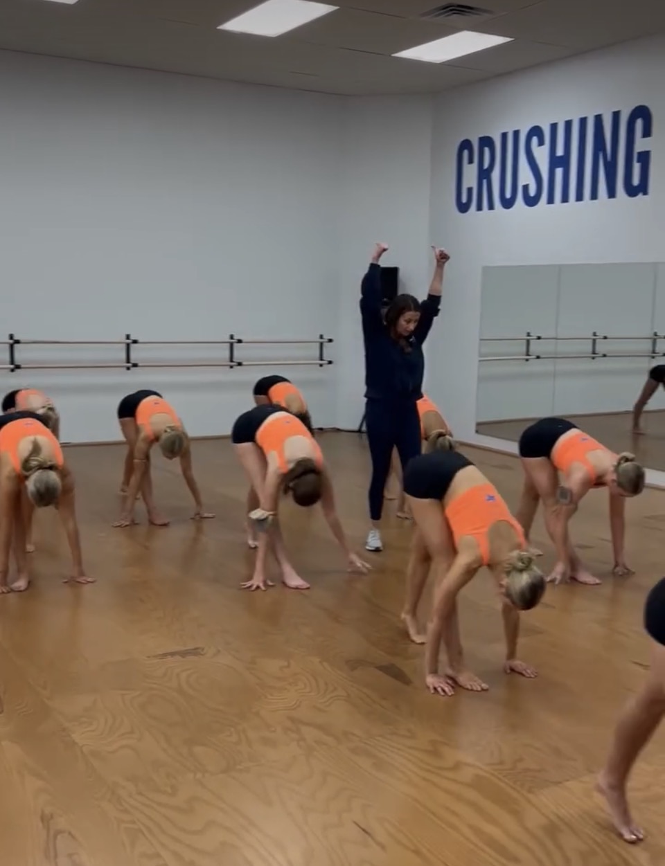 Group of young dancers in orange and black costumes bending forward in a dance studio, instructor with raised arms, word 'CRUSHING' on wall