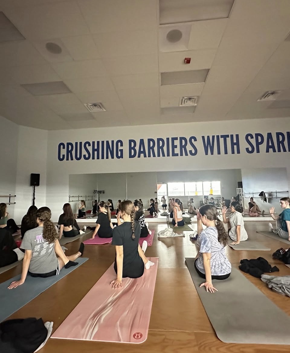 People participating in a yoga class in a spacious room with a large window and a banner reading 'CRUSHING BARRIERS WITH SPARK'.