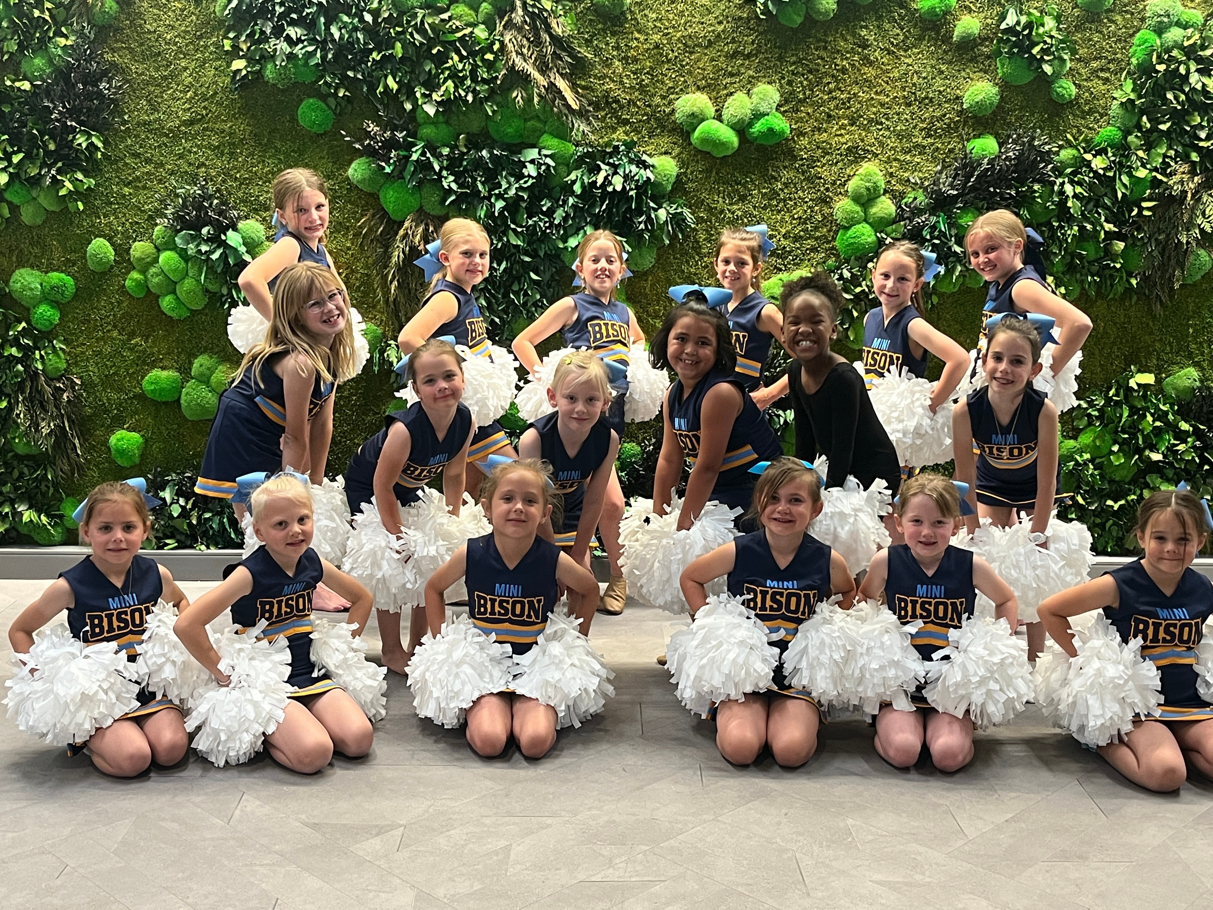 Group of young girls in cheerleading uniforms with white pom-poms, posing indoors in front of a green wall with plants.